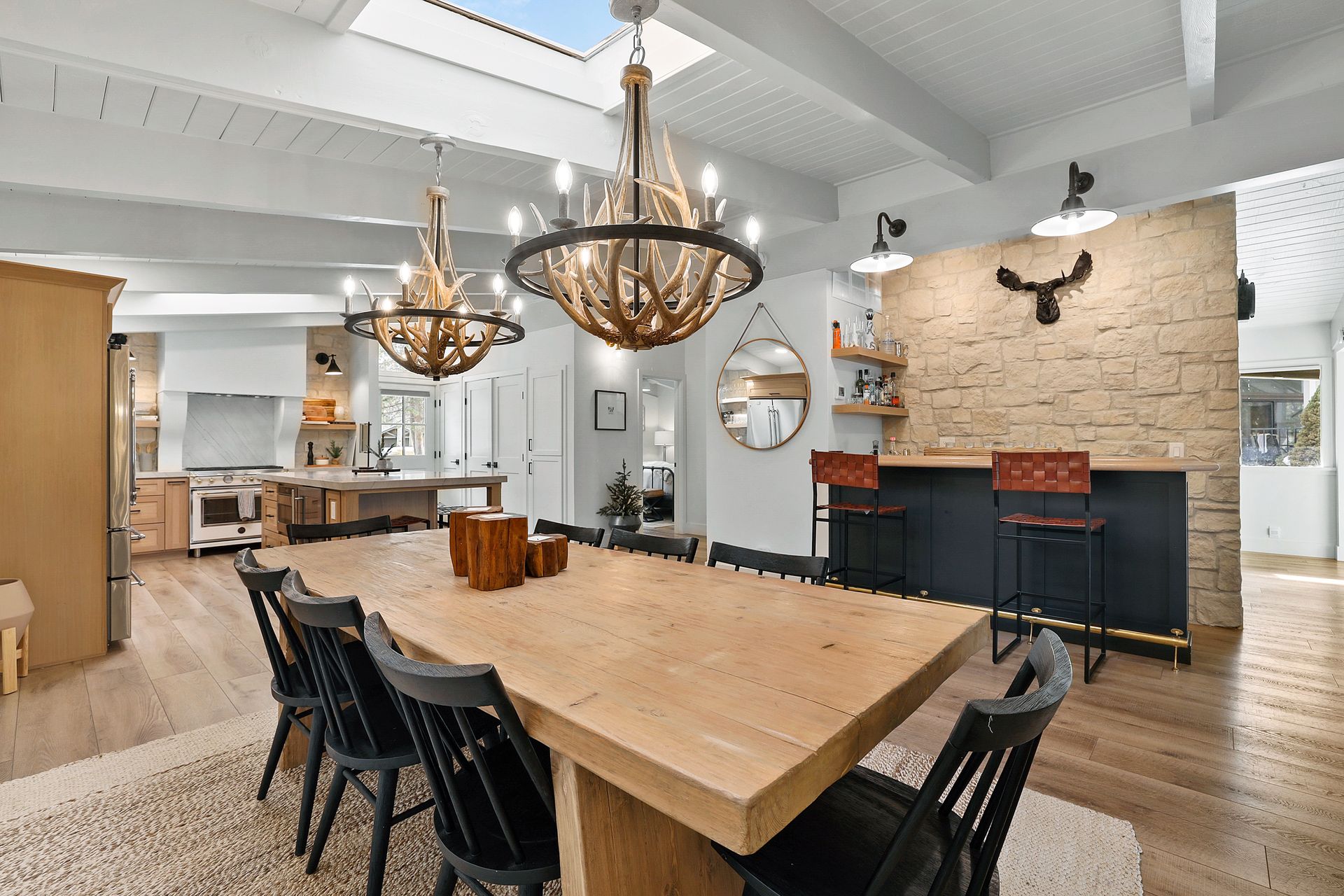 Modern dining room with a large wood table, black chairs, chandeliers, and a brick fireplace on the right.