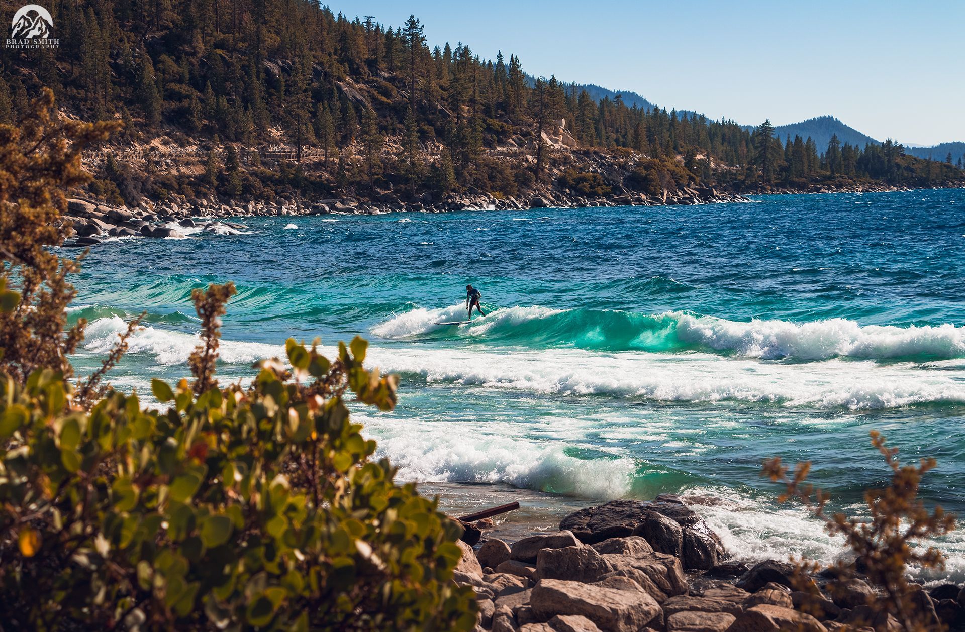 Rocky lakeshore with turquoise waves, pine-covered hills, and a person swimming in the distance