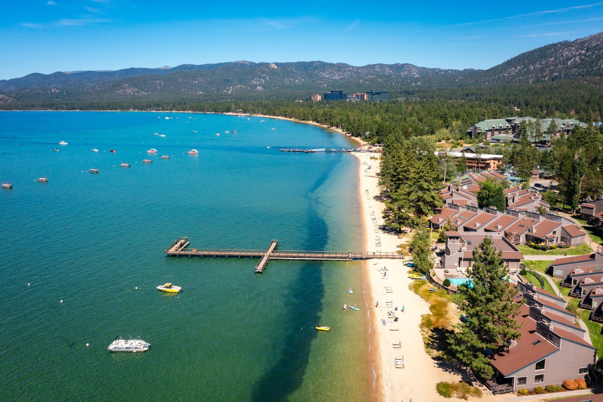 Aerial view of a turquoise lake, sandy beach, pier, and lakeside buildings with mountains in the distance