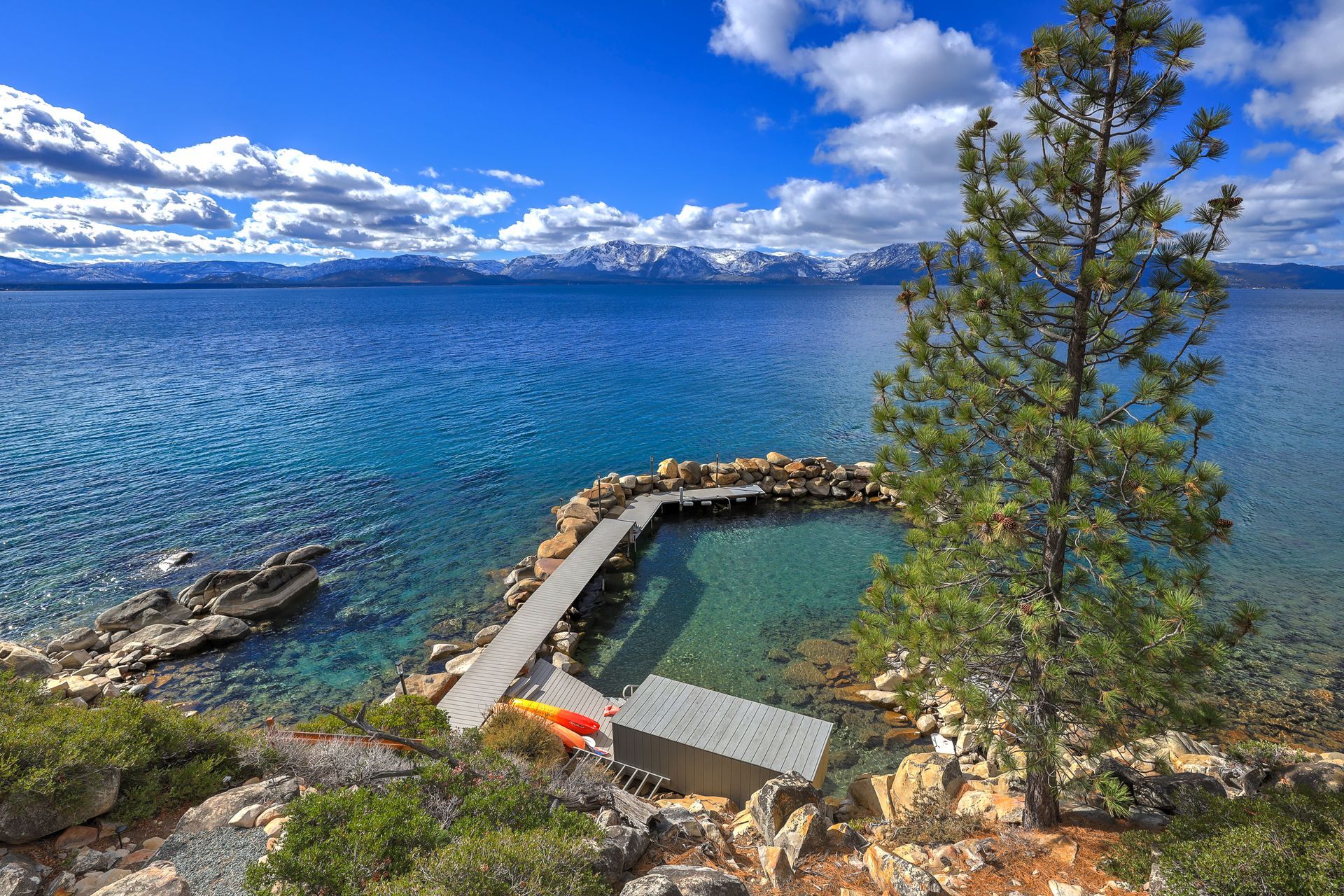 Rocky lakeshore with turquoise water, a small dock, pine trees, and snowy mountains under a bright blue sky
