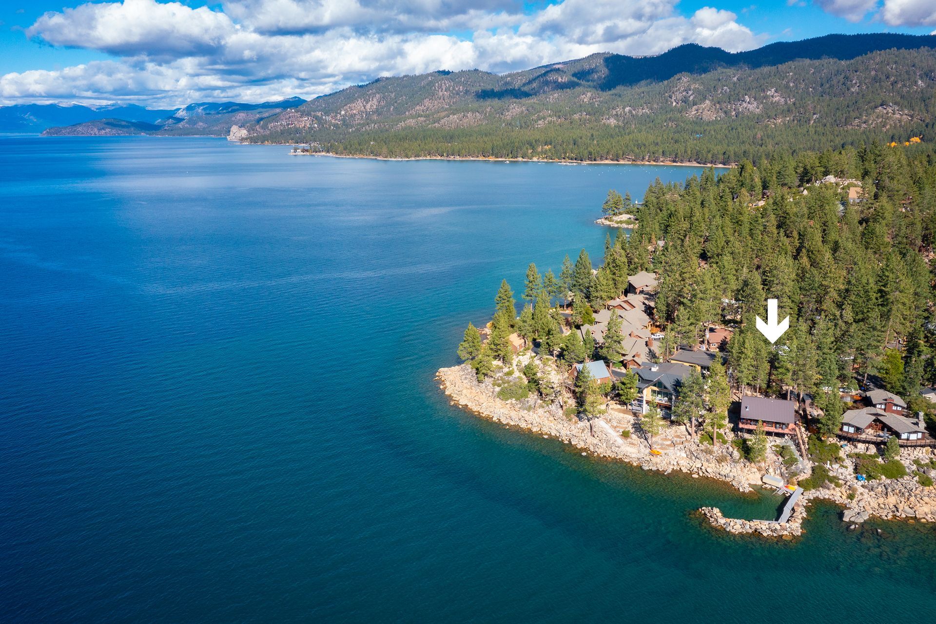Aerial view of a blue lake beside a wooded shoreline with houses and mountains in the distance