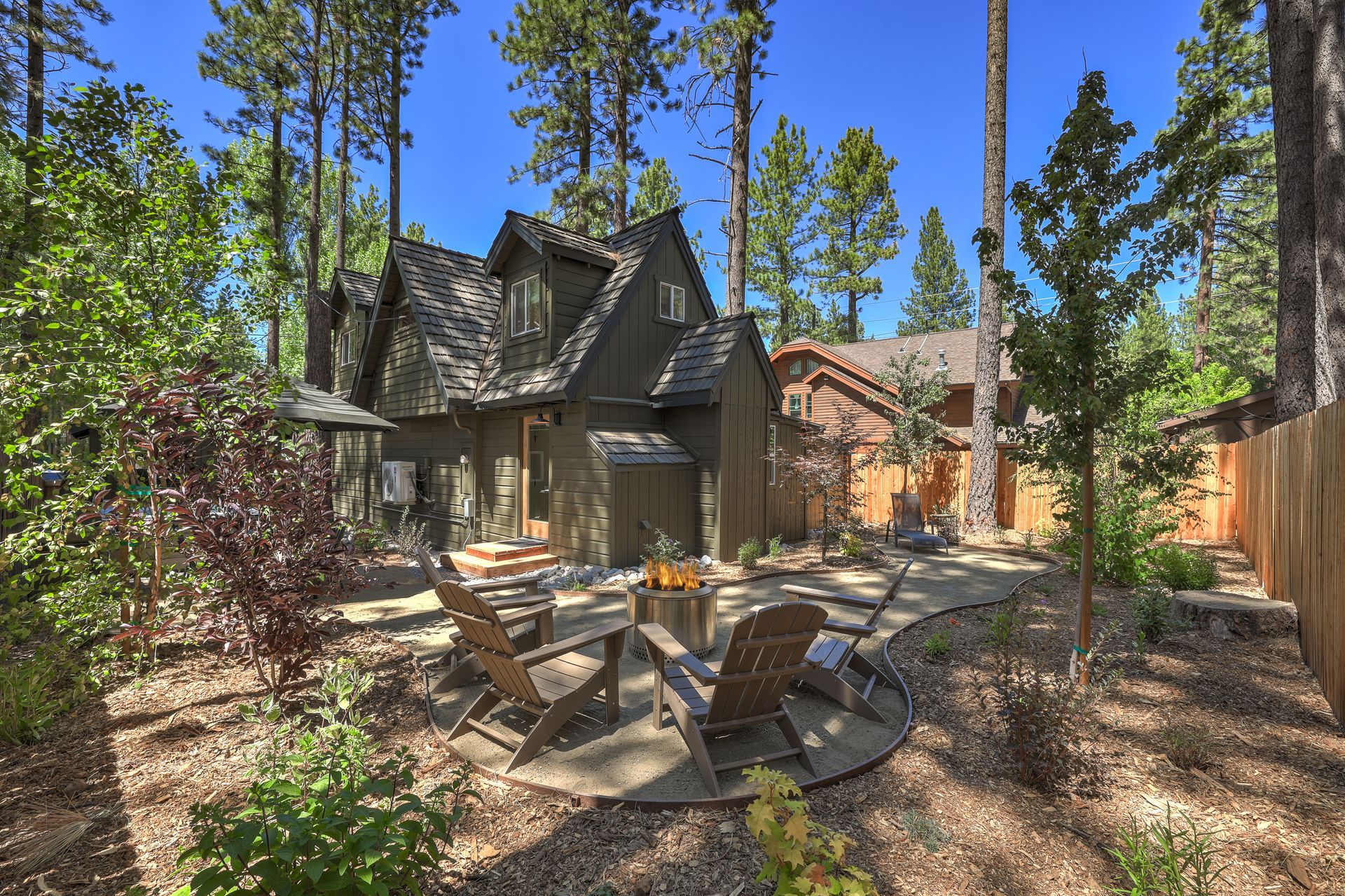 Wooded backyard patio with chairs and table beside a cabin-style house and tall pine trees