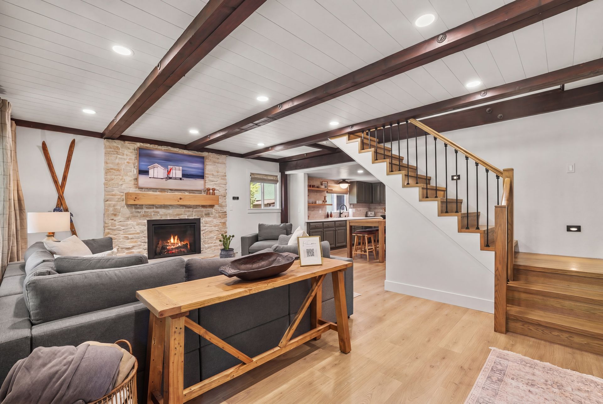 Cozy living room with gray sofa, fireplace, wood ceiling beams, and staircase leading to a kitchen.