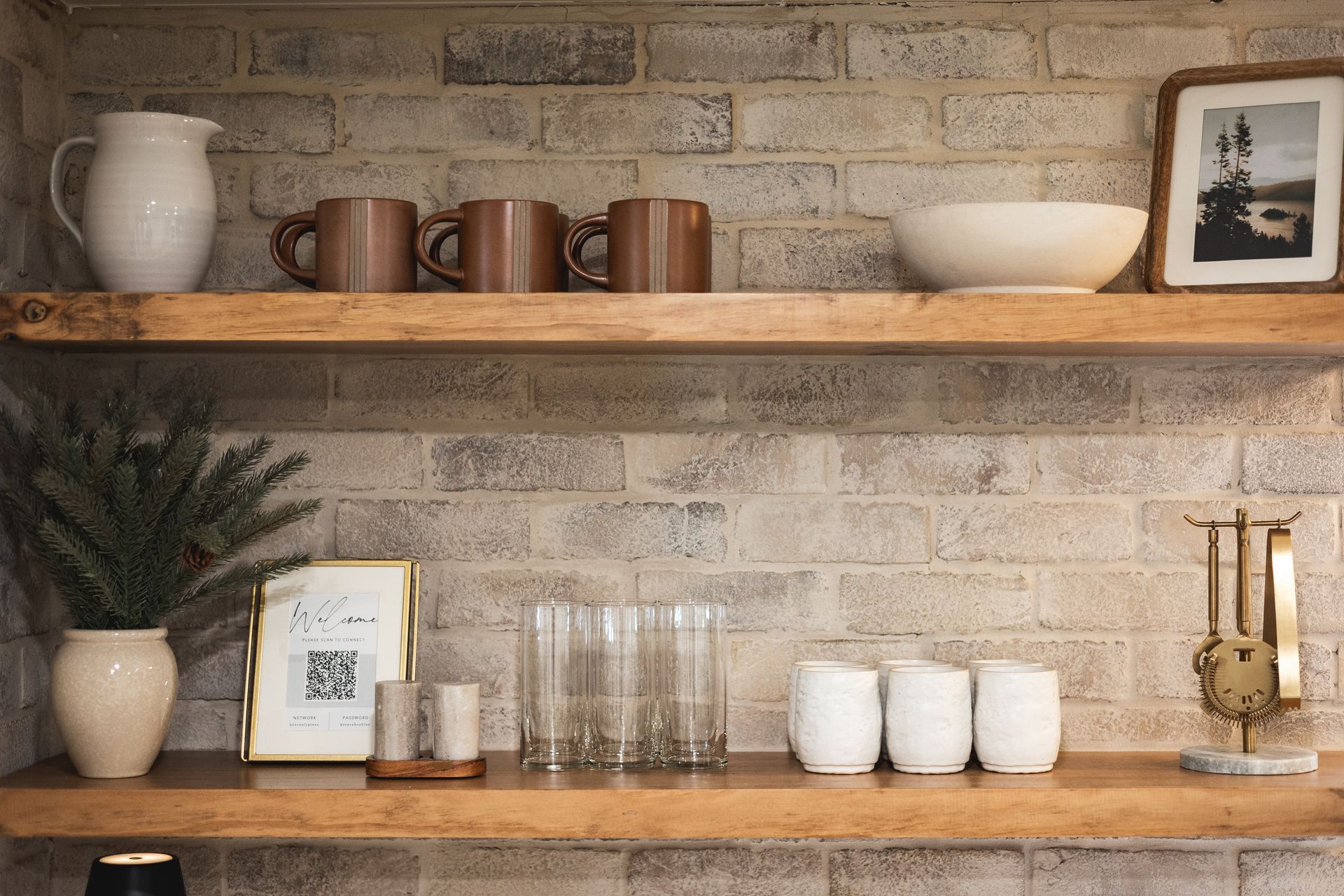 Rustic kitchen shelves with mugs, bowls, glasses, candles, a plant, and framed art against a brick wall