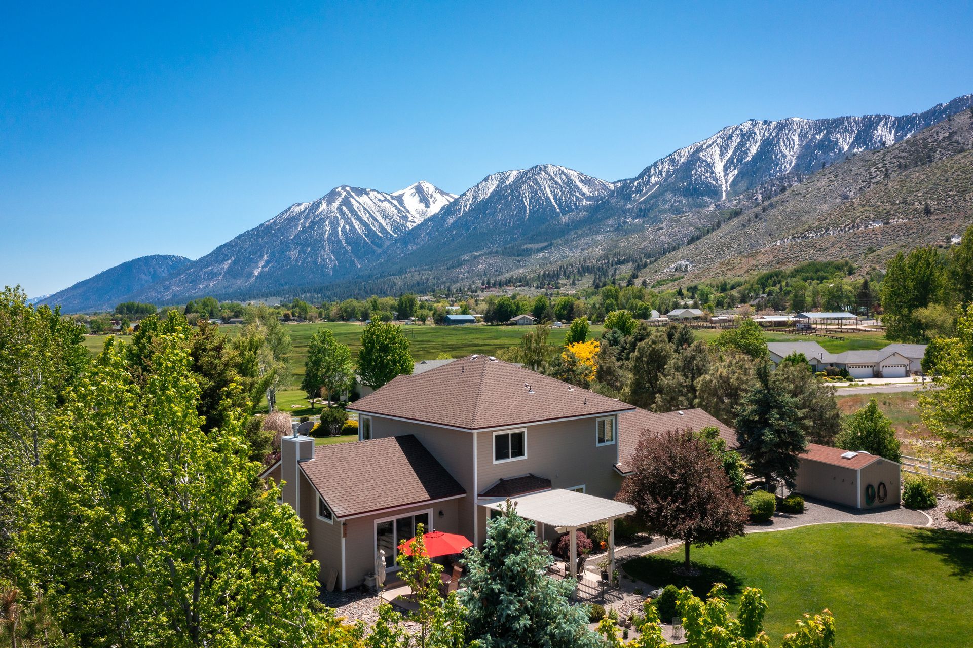 Suburban home with a backyard, green trees, and snow-capped mountains under a clear blue sky