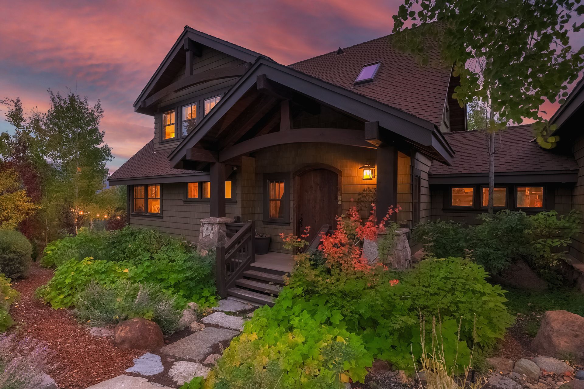 Cozy dark-wood cabin at dusk with glowing windows, stone steps, and lush garden under a pink sky