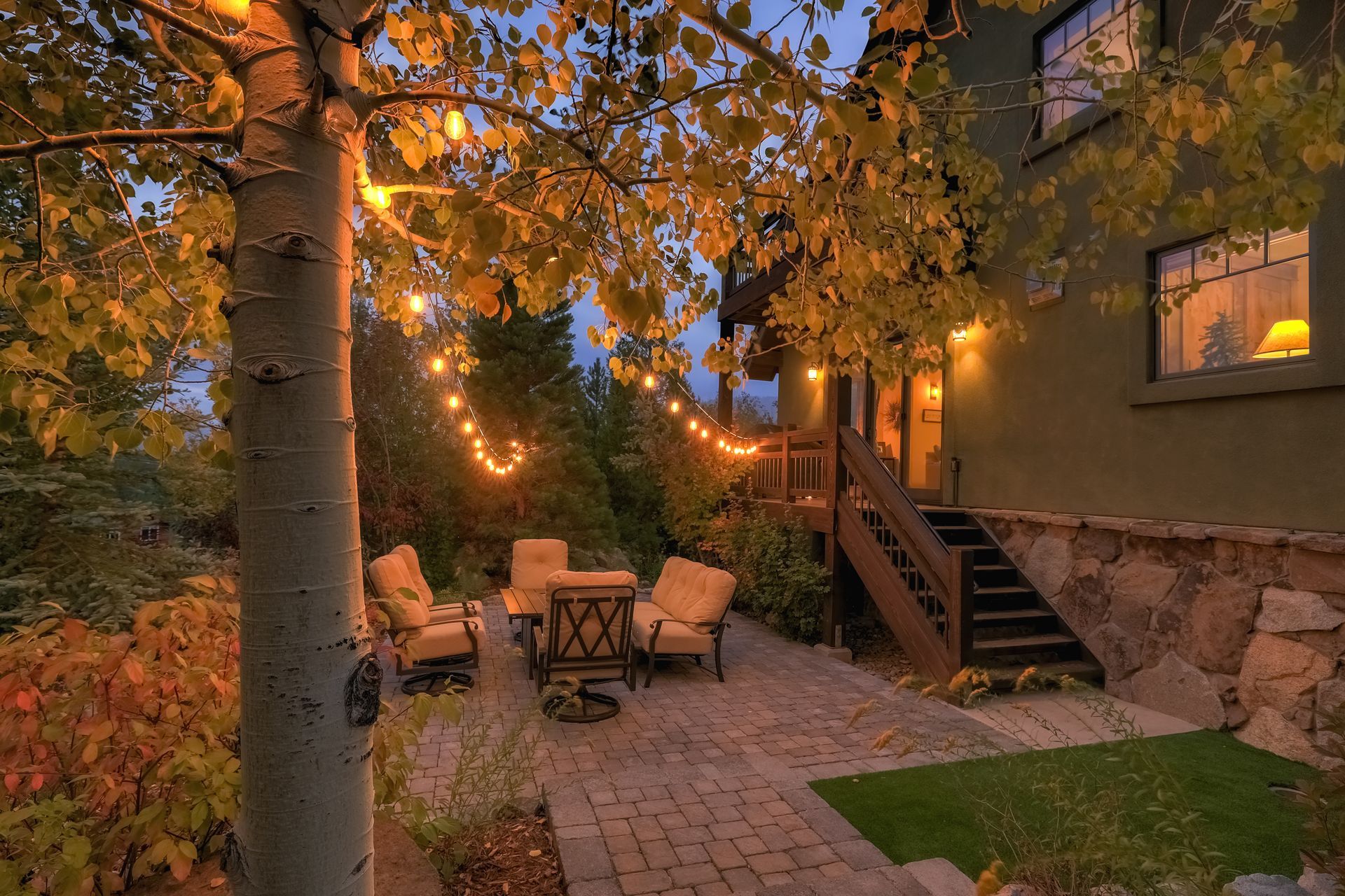 Cozy stone patio with string lights, outdoor seating, and autumn trees beside a rustic house