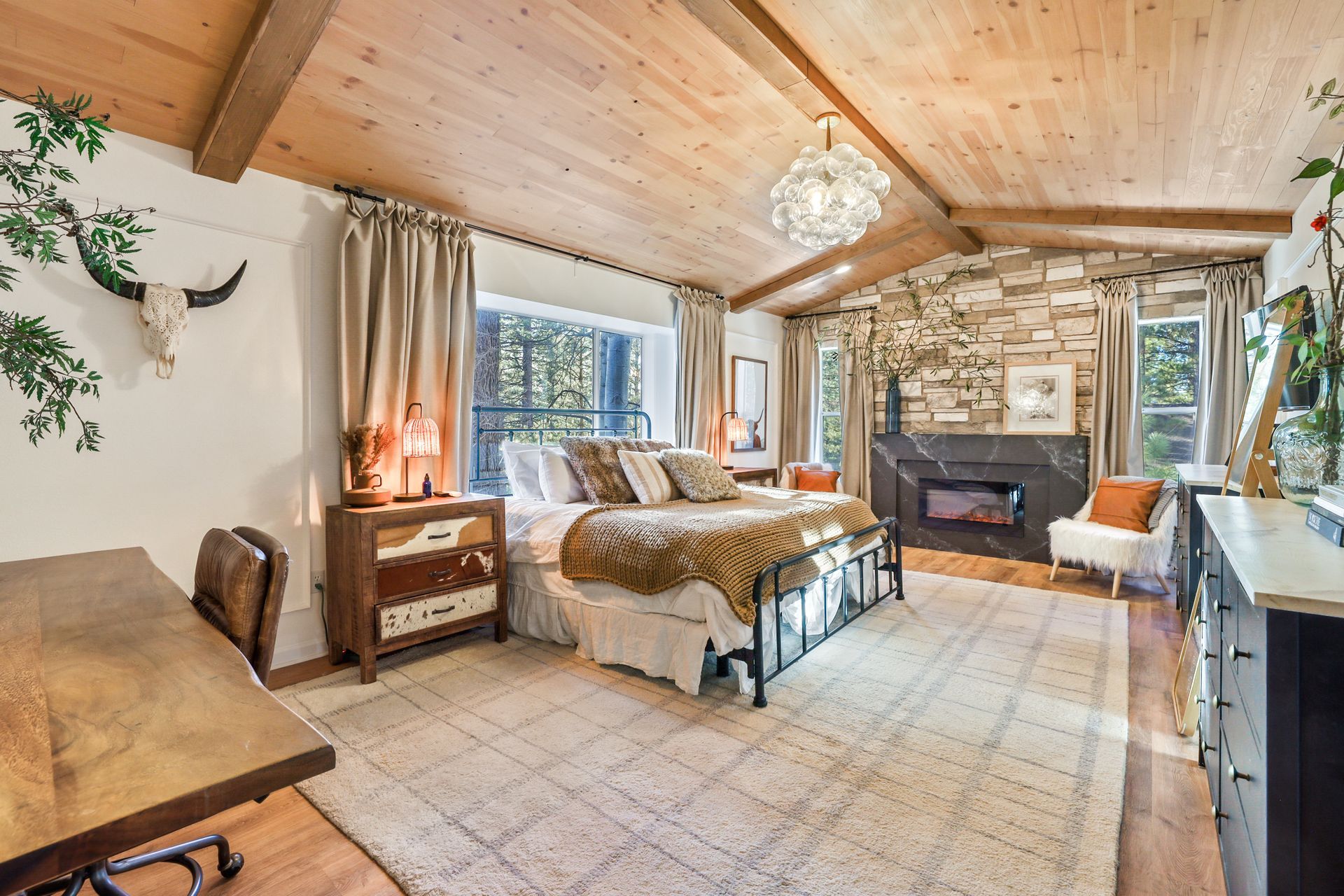 Cozy rustic bedroom with wood ceiling, stone fireplace, bed, window, and tiled floor