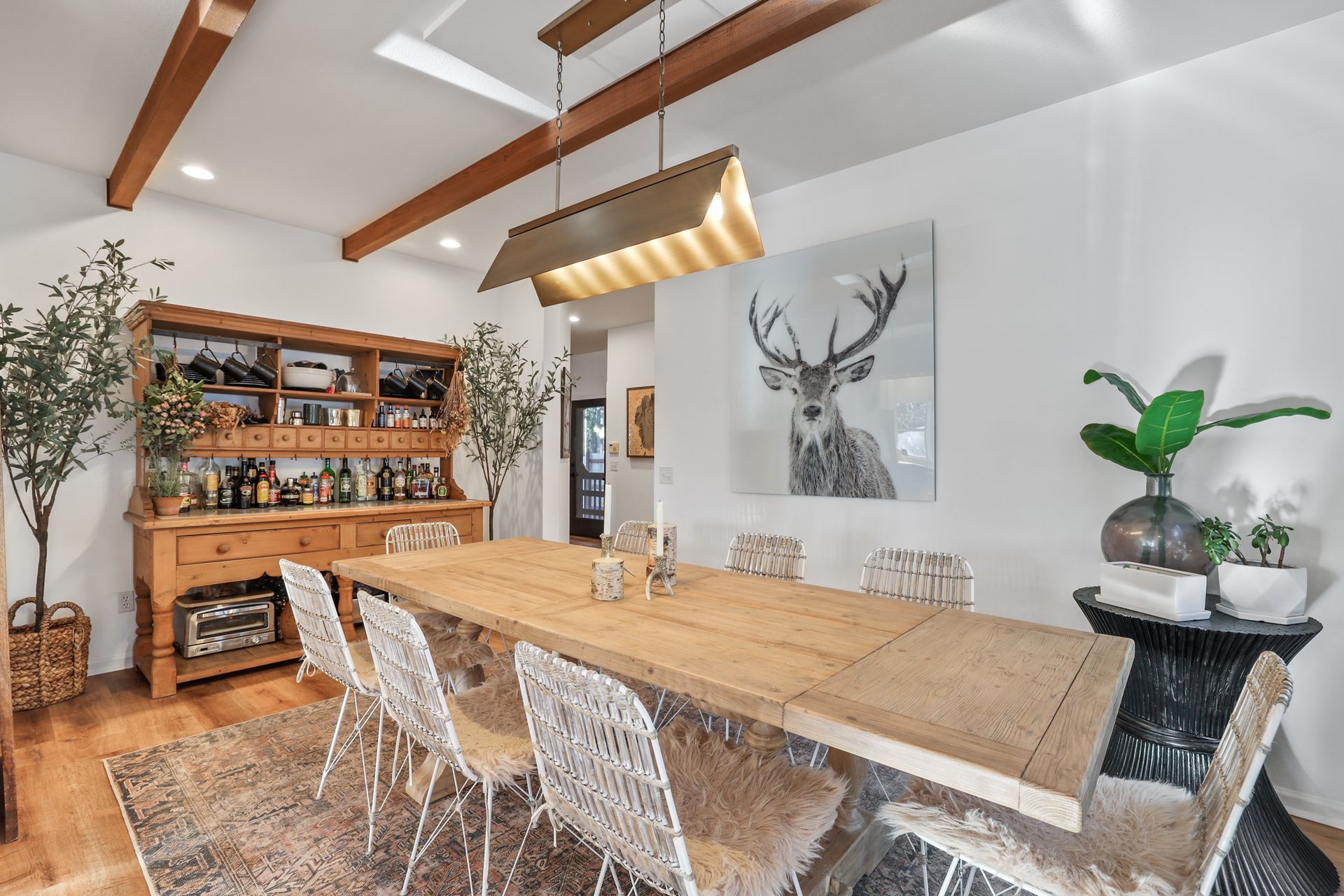 Modern dining room with a long wooden table, white chairs, deer wall art, and a shelving unit under exposed beams
