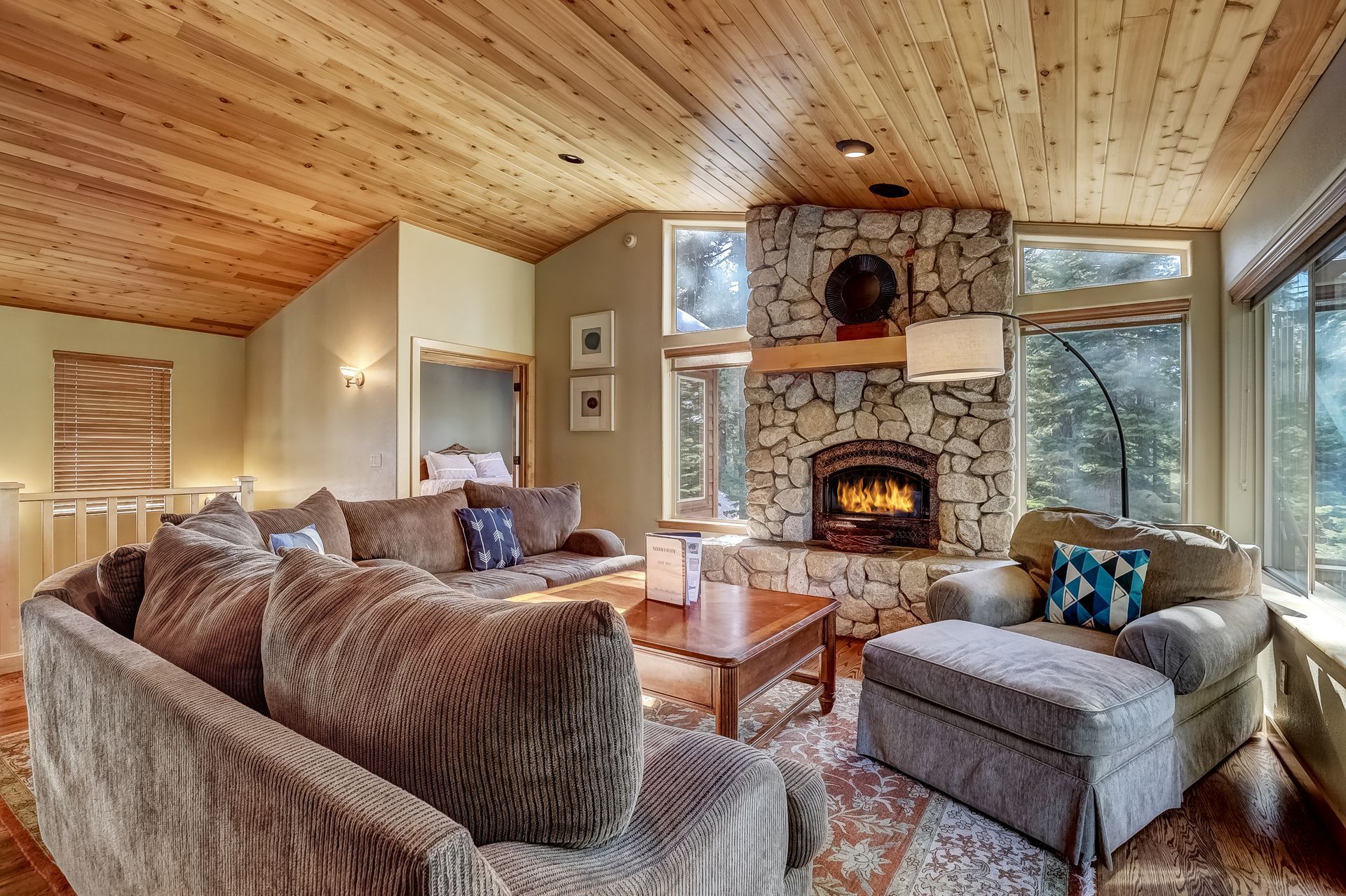 Cozy living room with stone fireplace, wood ceiling, large sectional sofa, and forest-view windows