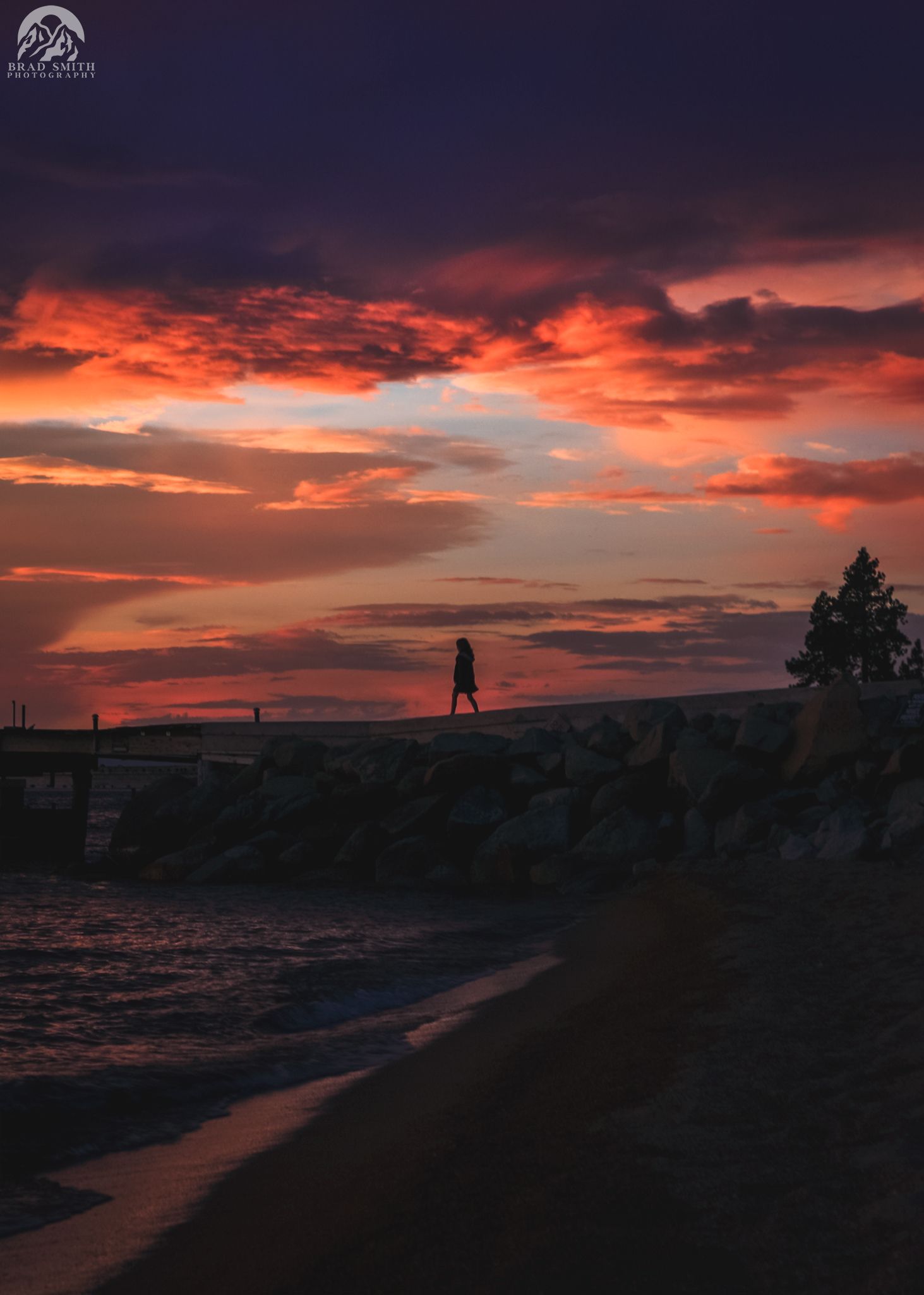 Sunset over a rocky shoreline with dramatic red-orange clouds and a silhouetted person on the horizon