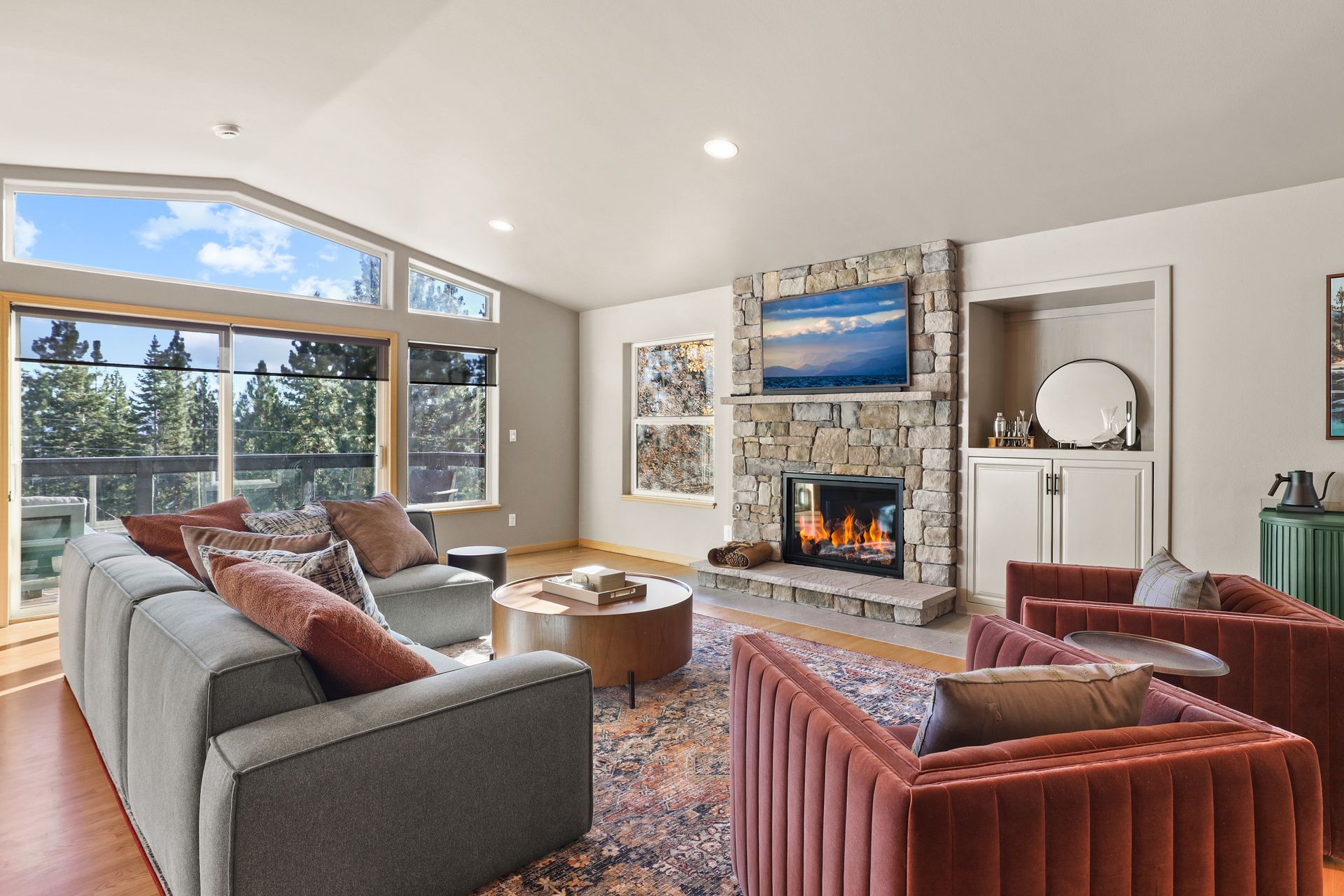 Bright living room with large windows, gray sofa, red chairs, fireplace, and mountain view