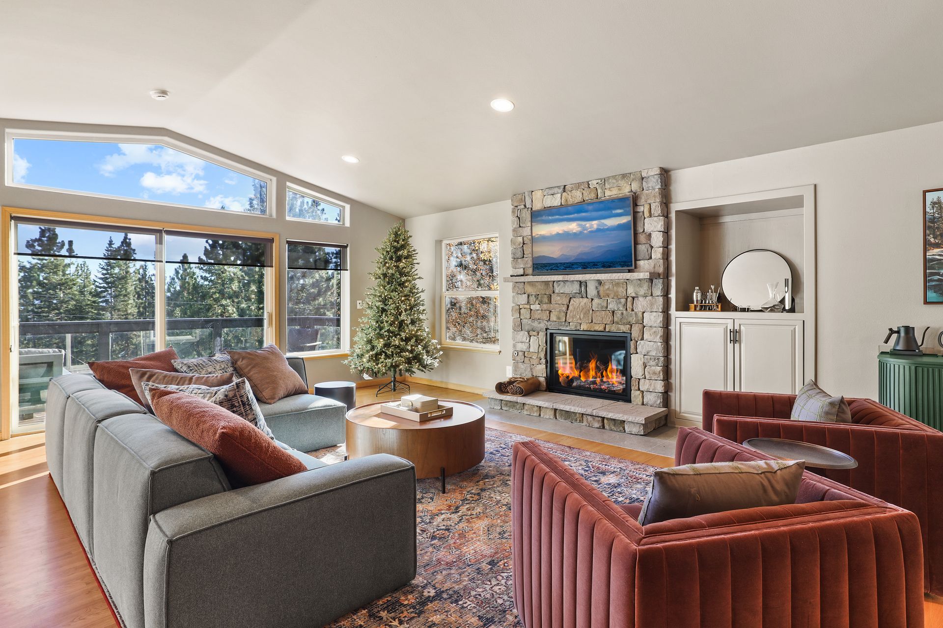 Bright living room with large windows, gray sofa, red chair, fireplace, and TV above the mantel