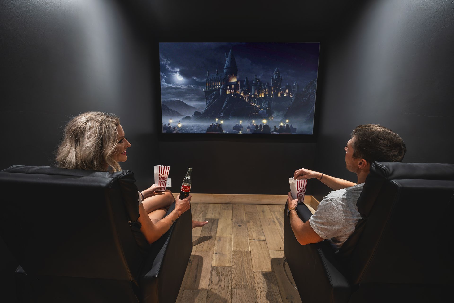 Two people sit in a dark home theater watching a movie on a large screen, drinks in hand.