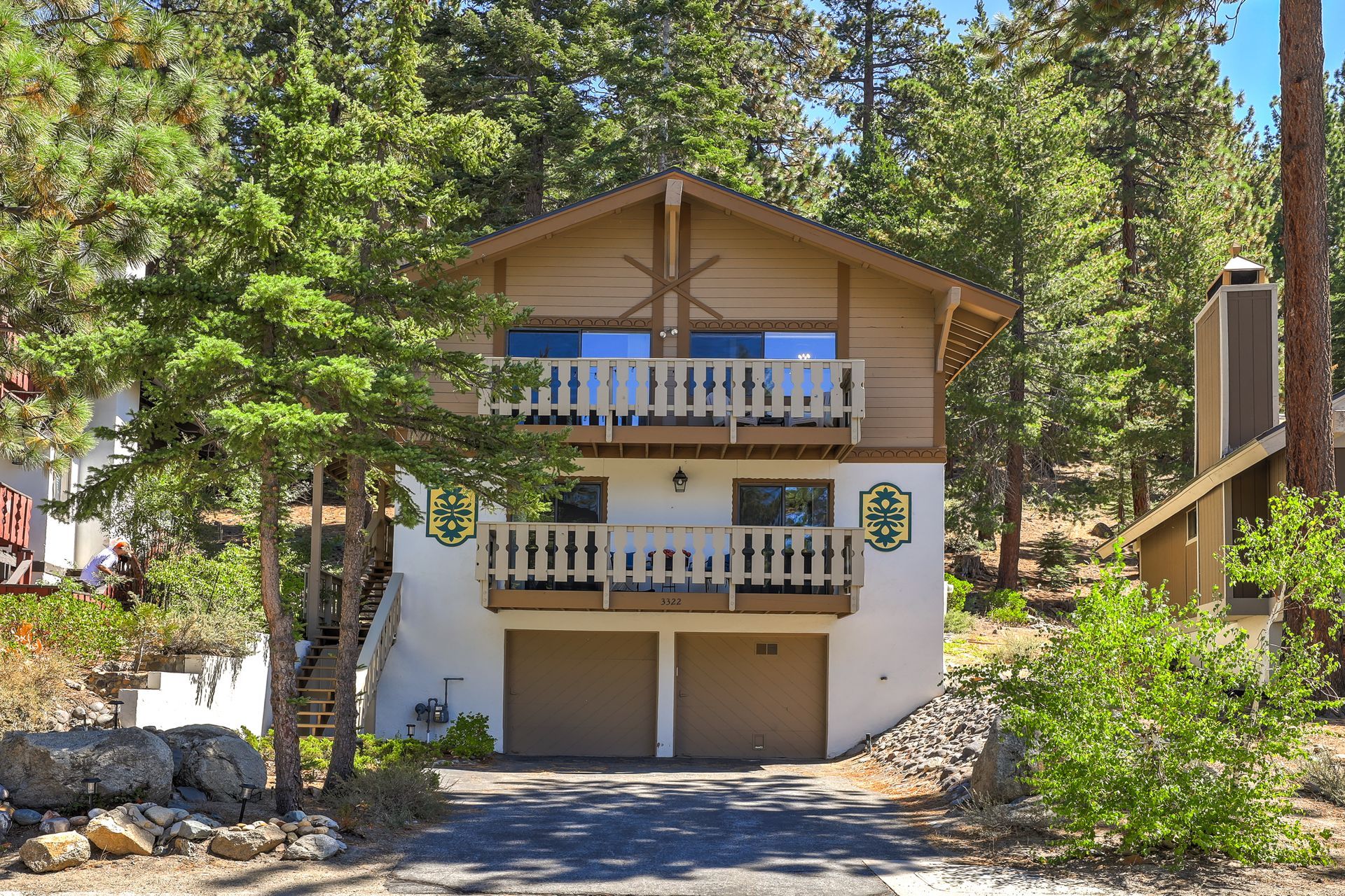 Two-story mountain cabin with balconies and garage, surrounded by tall pine trees and greenery