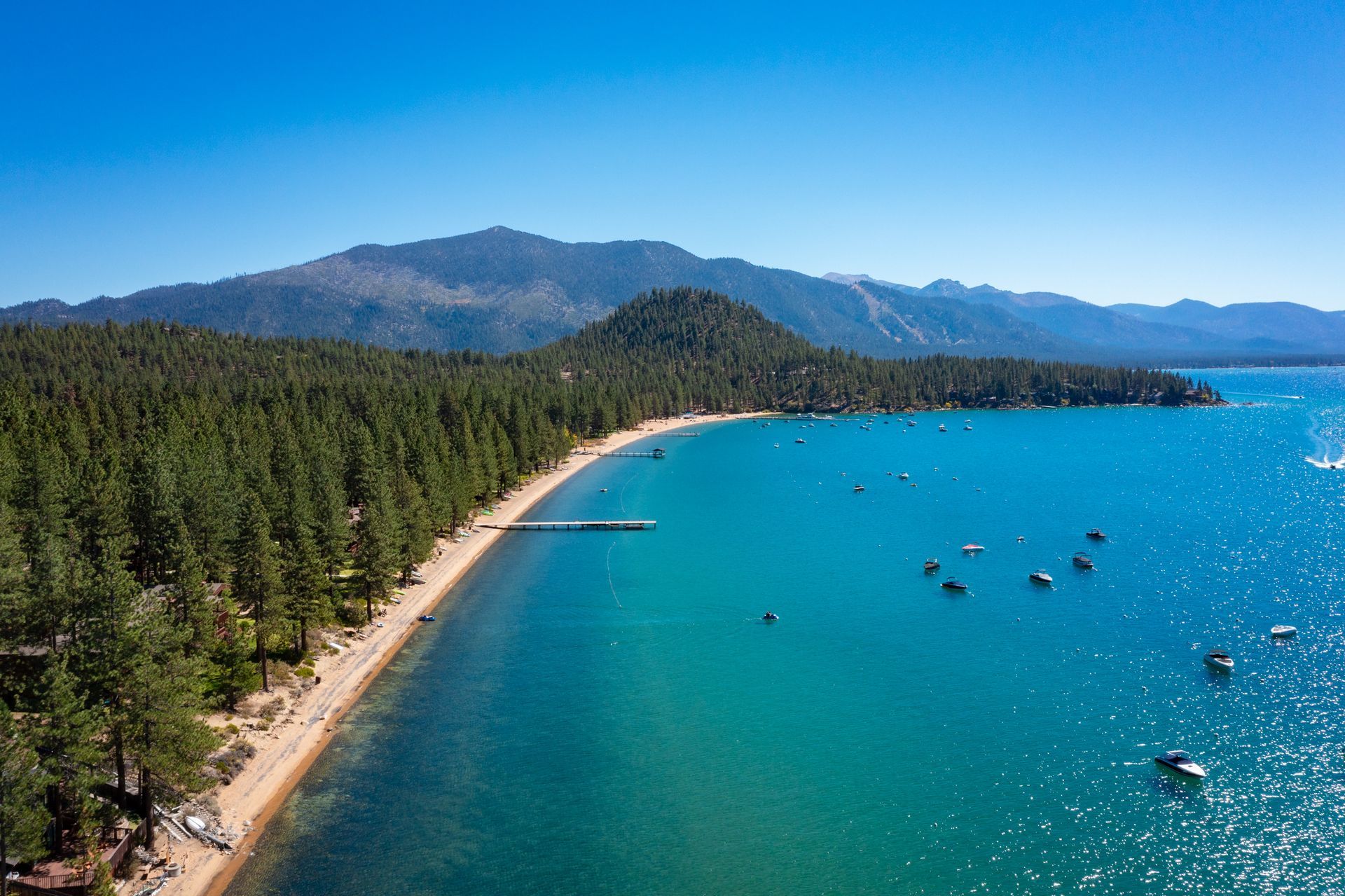 Aerial view of a crescent beach, pine forest, and turquoise bay with small boats and distant mountains