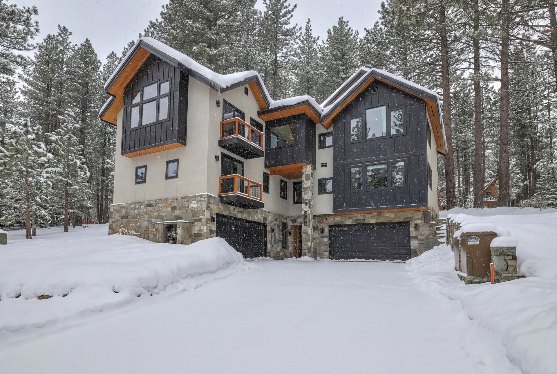 Snow-covered modern cabin with stone base and dark siding in a pine forest