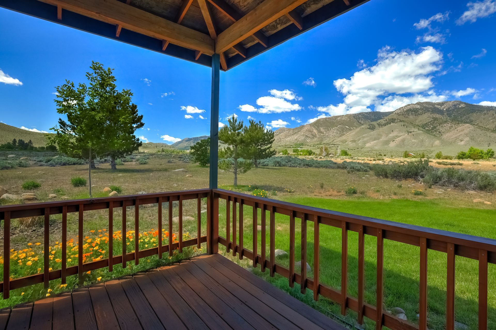 Wooden porch overlooking green fields, mountains, and a bright blue sky with clouds.
