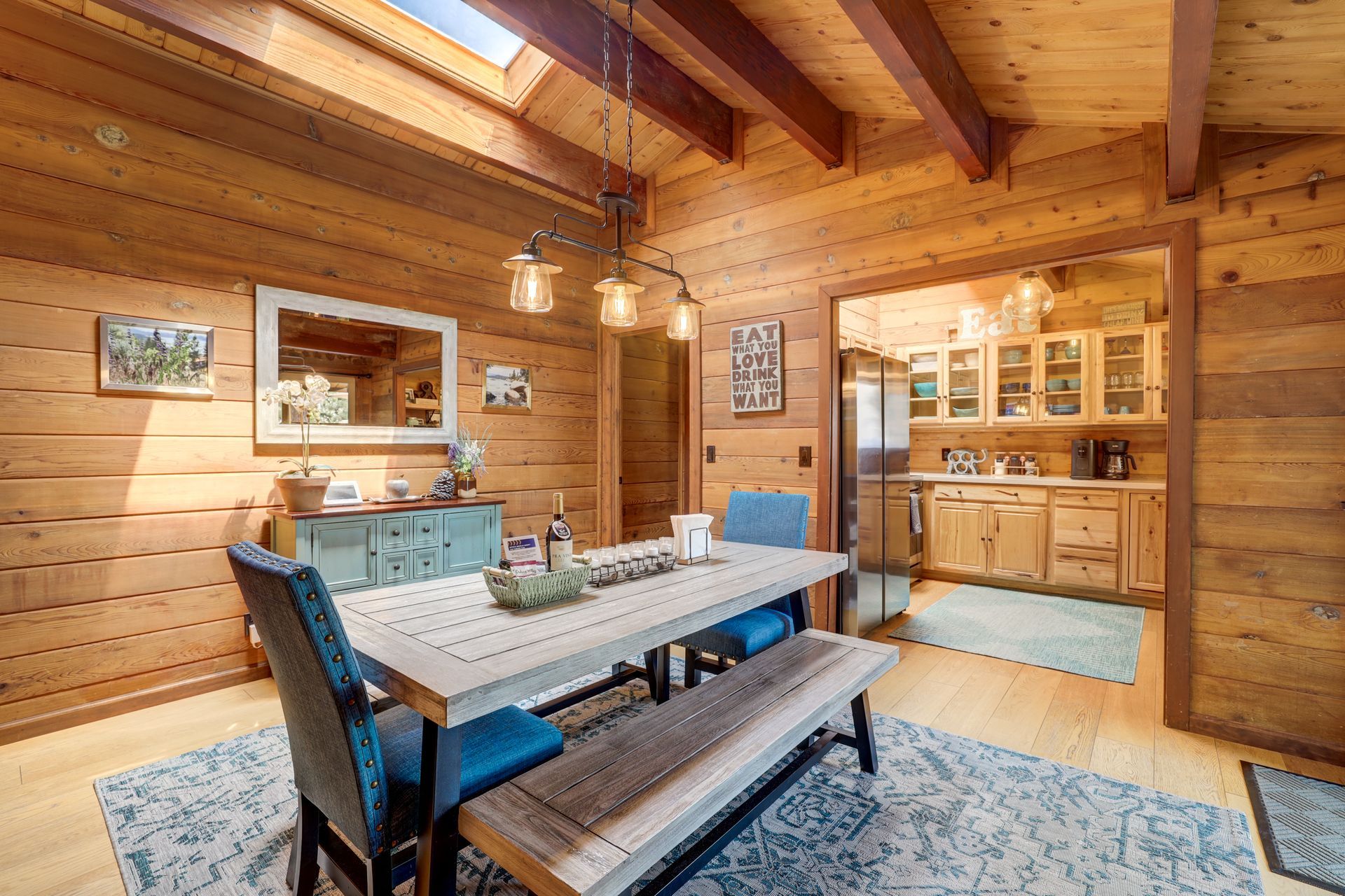Rustic dining room with wooden walls, blue chairs, and a long table beside a kitchen pass-through.