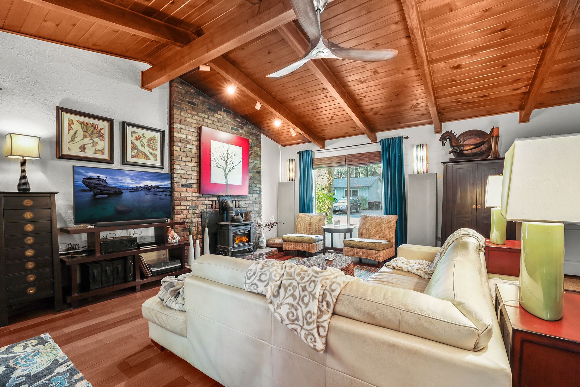 Cozy living room with wood-beamed ceiling, white sofa, TV, fireplace, and large windows overlooking water.