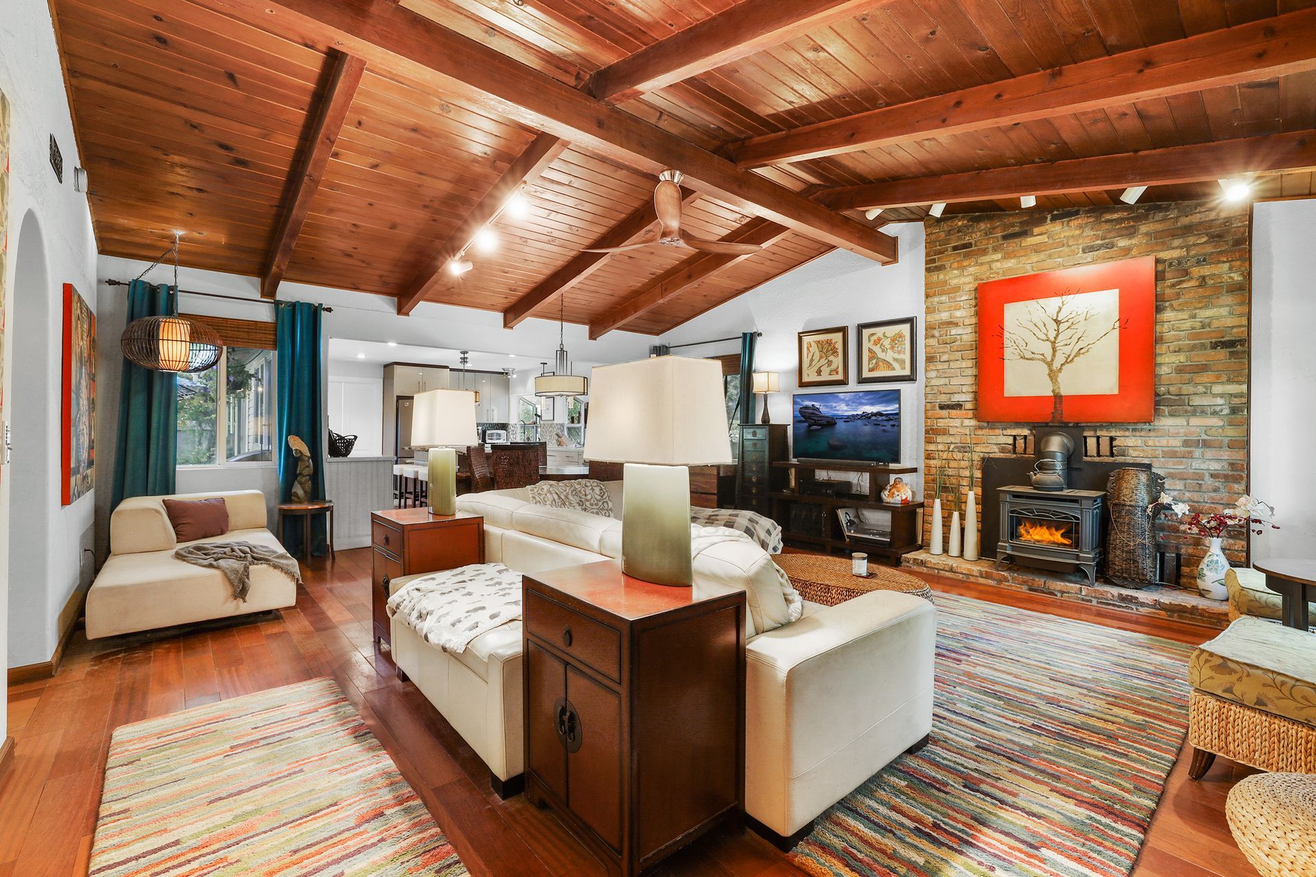 Warm living room with wood-beamed ceiling, stone fireplace, white sofa, and colorful rugs.