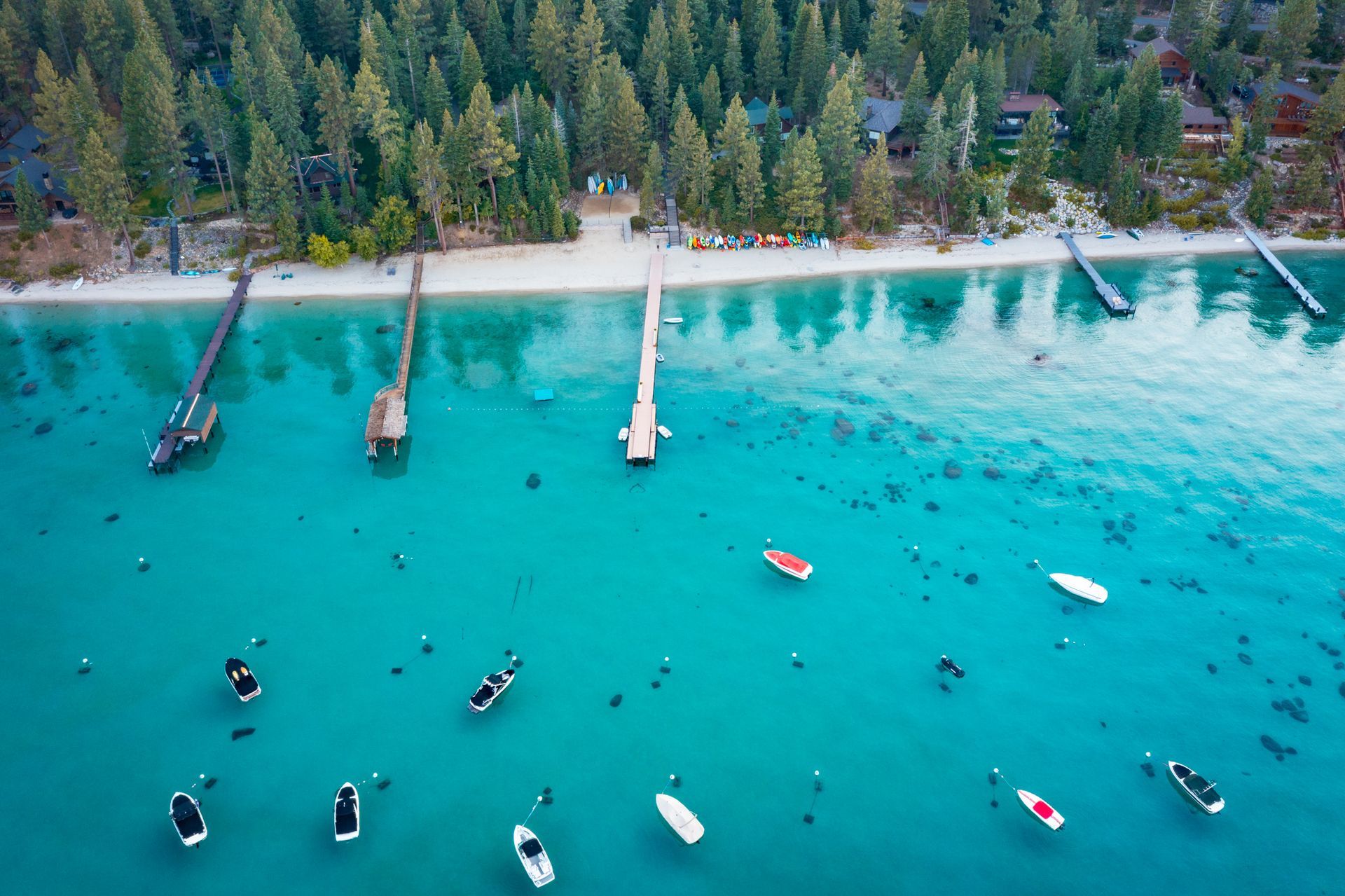 Aerial view of turquoise water with boats near a sandy beach and pine forest shoreline