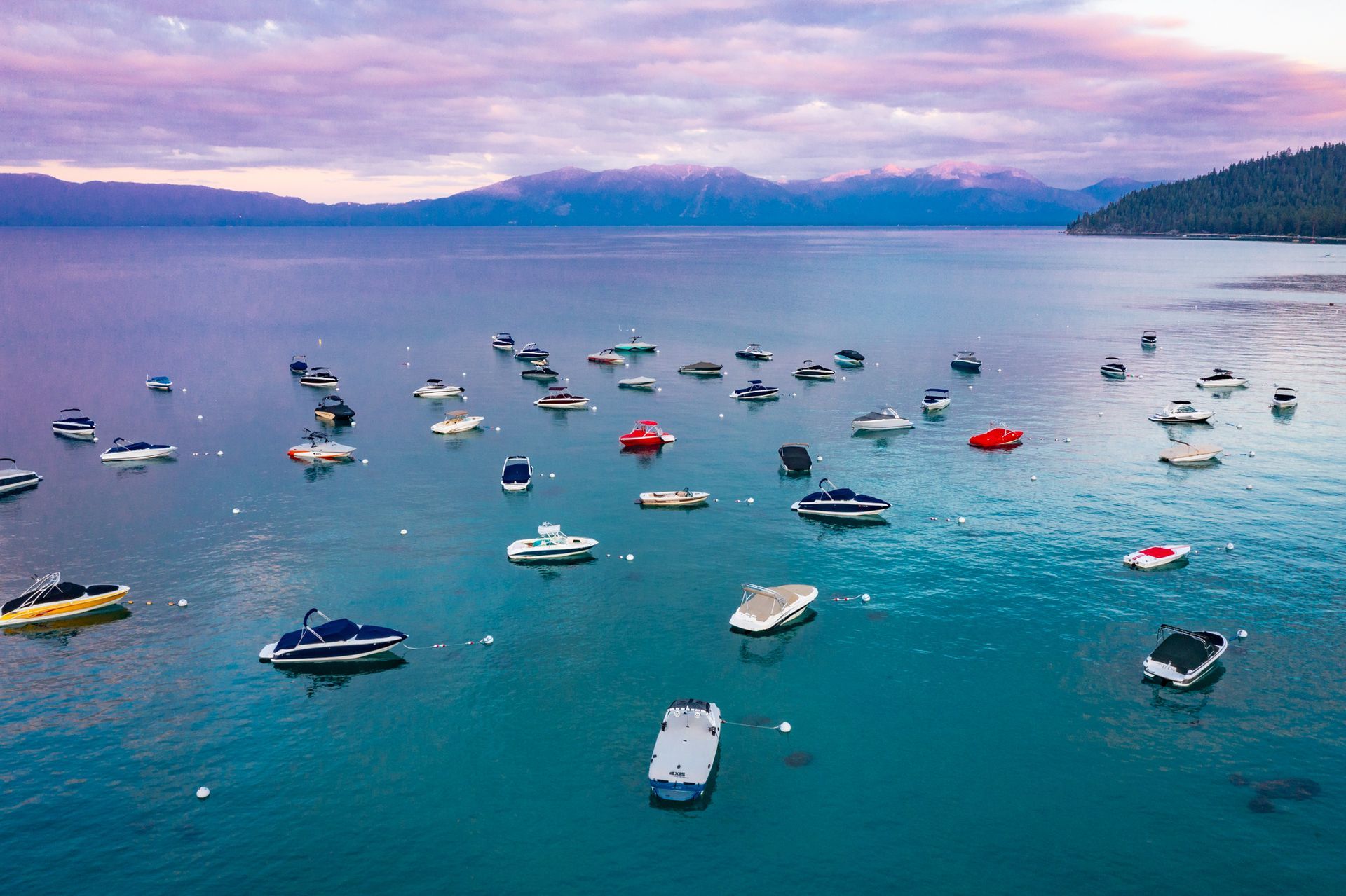 Many boats anchored on a calm blue lake at sunset, with mountains in the distance.