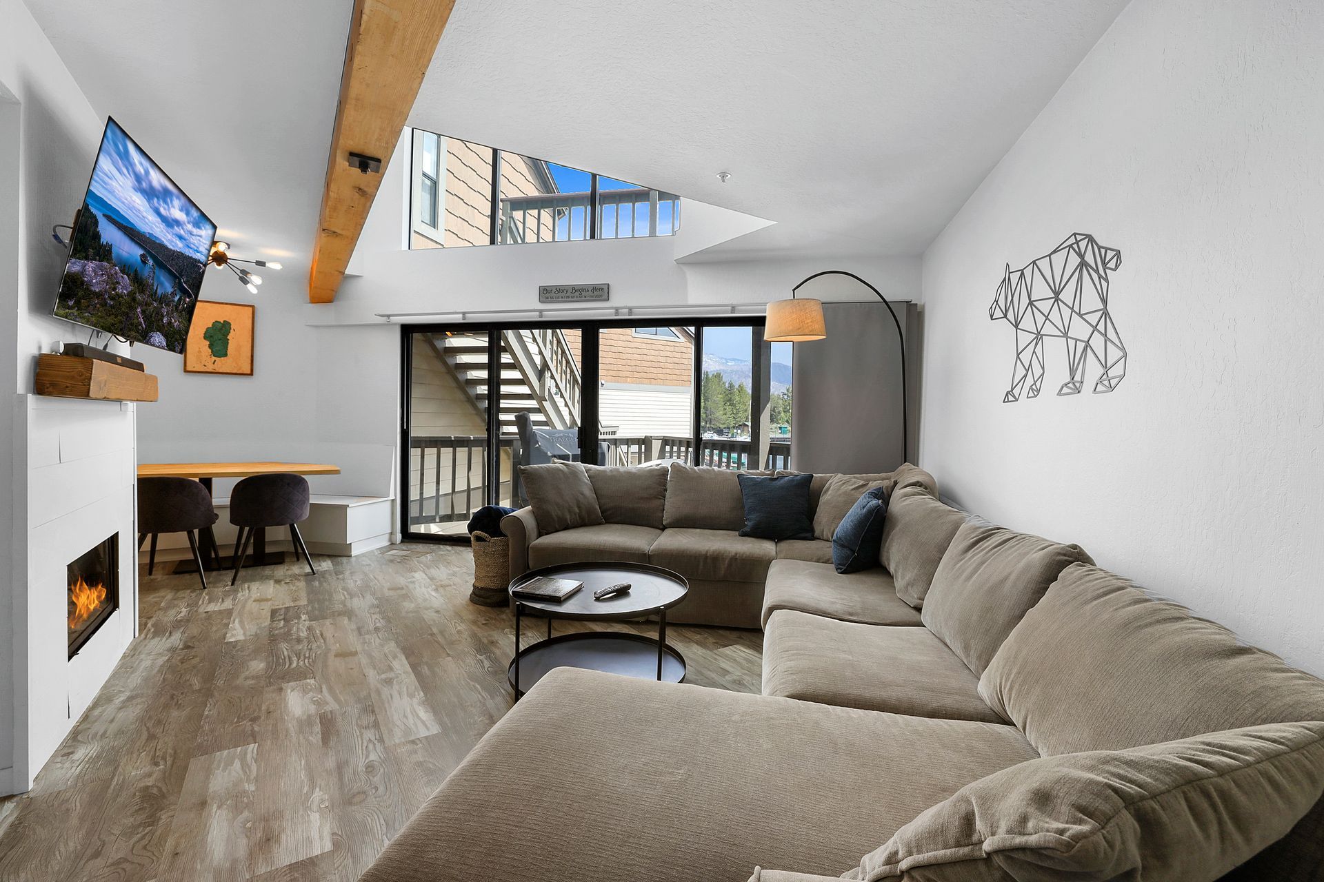 Modern loft living room with sectional sofa, wall-mounted TV, fireplace, and staircase under skylight.