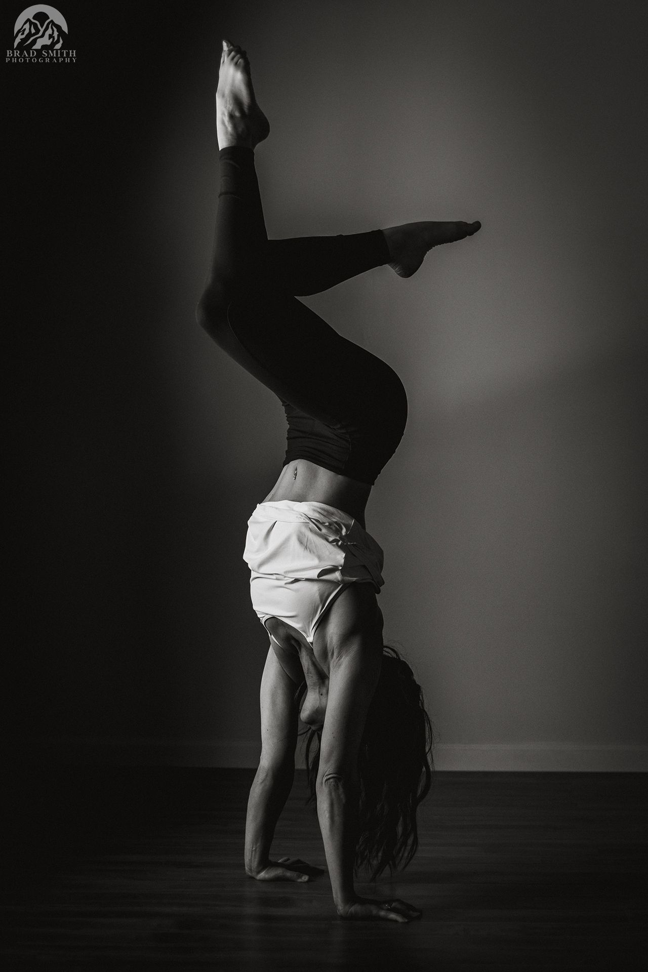 Black-and-white photo of a dancer balancing in a one-handed handstand against a plain wall.