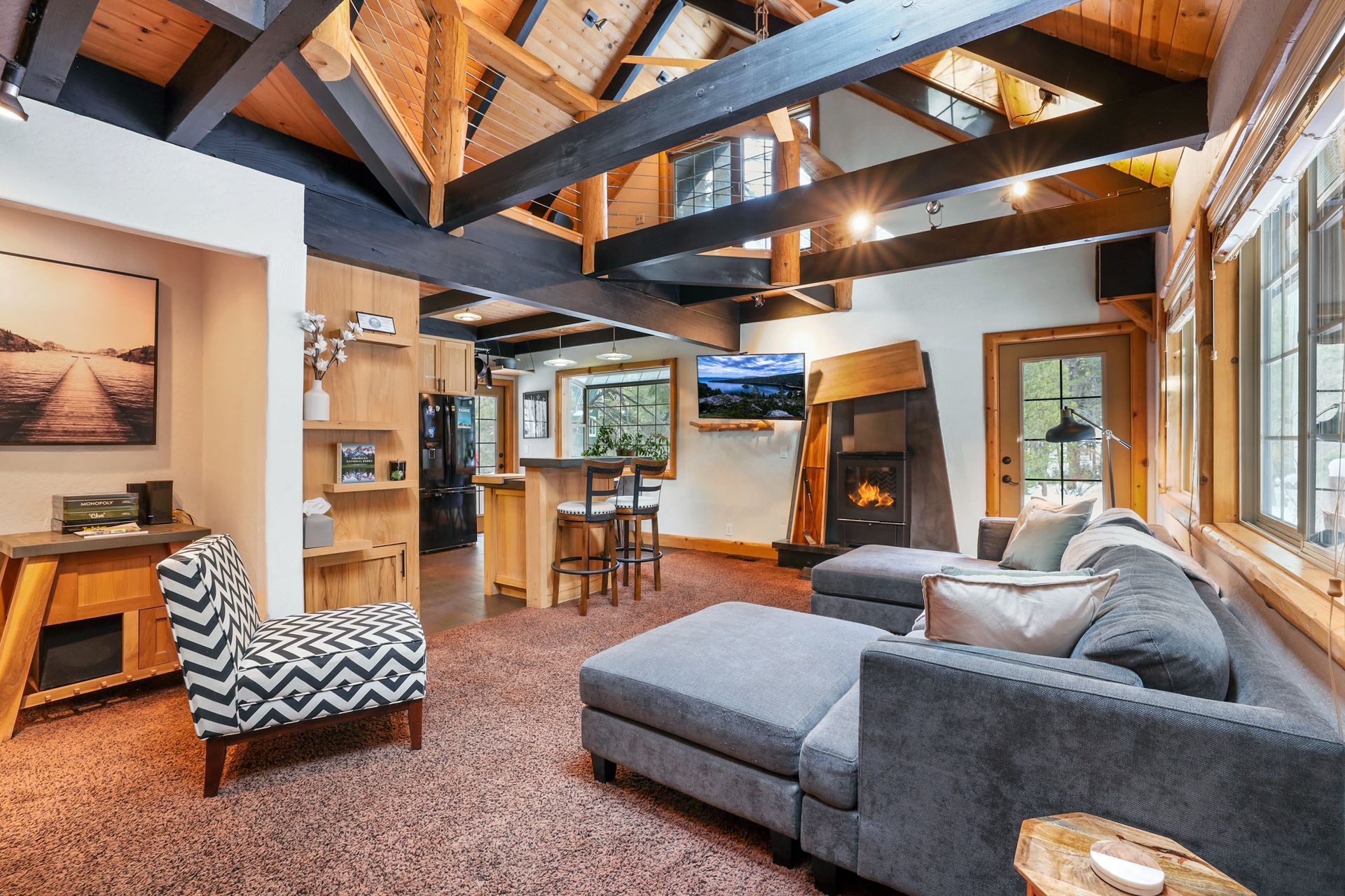 Cozy living room with gray sectional, wooden beams, and large windows overlooking a kitchen area.