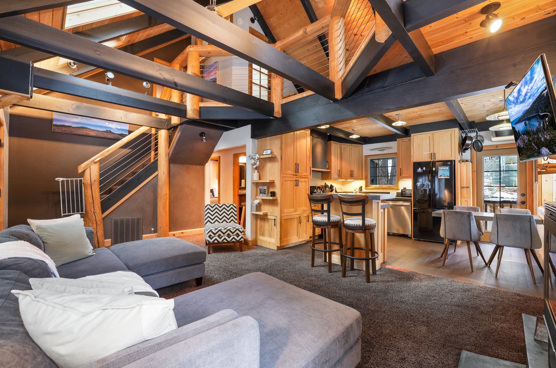 Cozy open-plan living room with exposed beams, gray sofa, and kitchen/dining area in warm wood tones