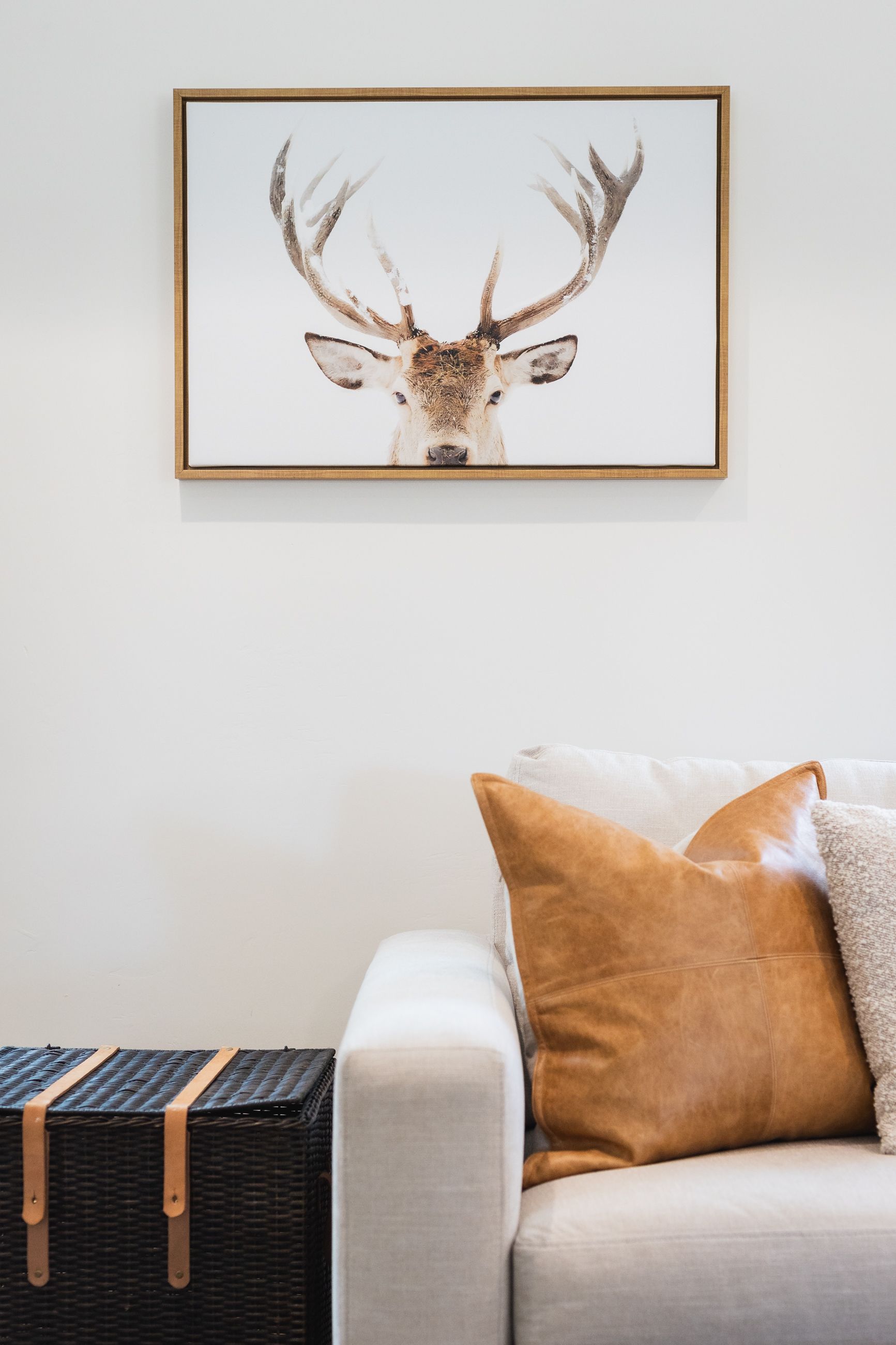 Framed deer print above a beige sofa with mustard and cream pillows beside a woven chest.
