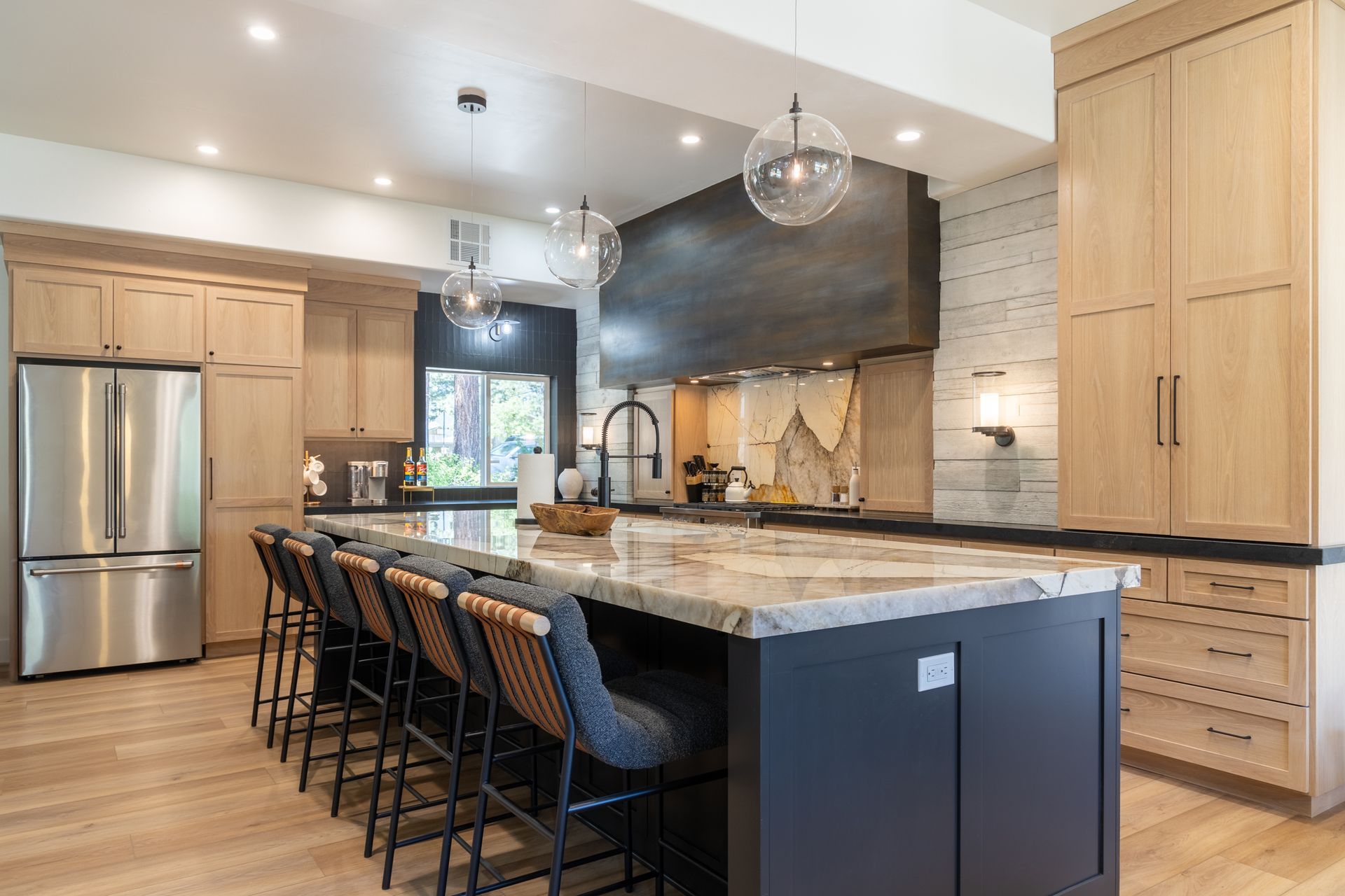 Modern kitchen with a large island, black stools, stainless steel fridge, and light wood cabinets.