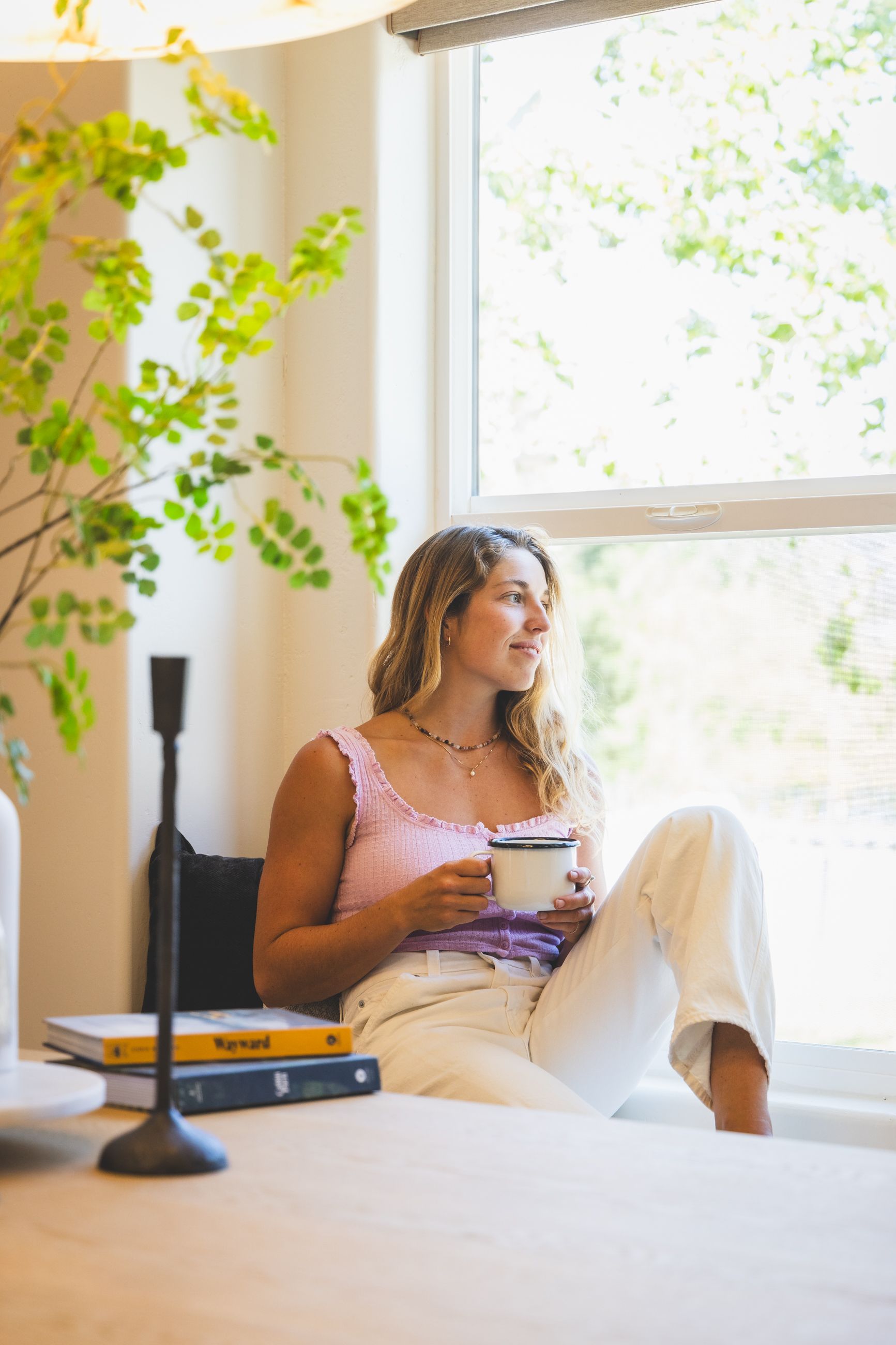 Person sitting on a windowsill holding a mug, surrounded by plants in a sunlit room.