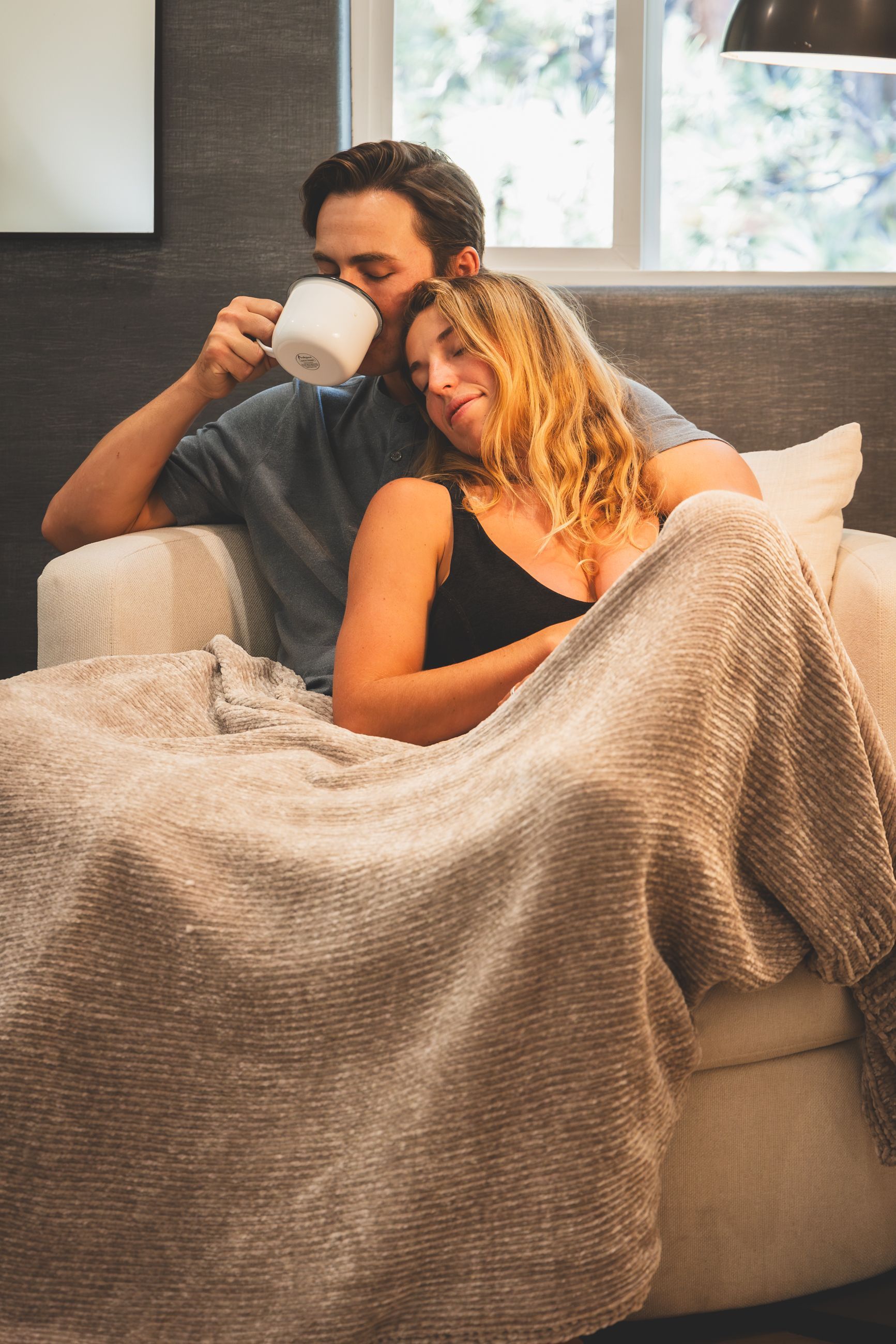 Couple relaxing under a beige blanket on a couch, one sipping from a white mug in a cozy room.