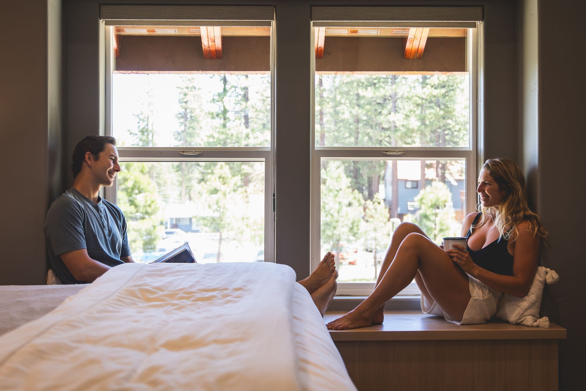 Two people relaxing in a bedroom by large windows, one sitting on the bed and one on a bench.