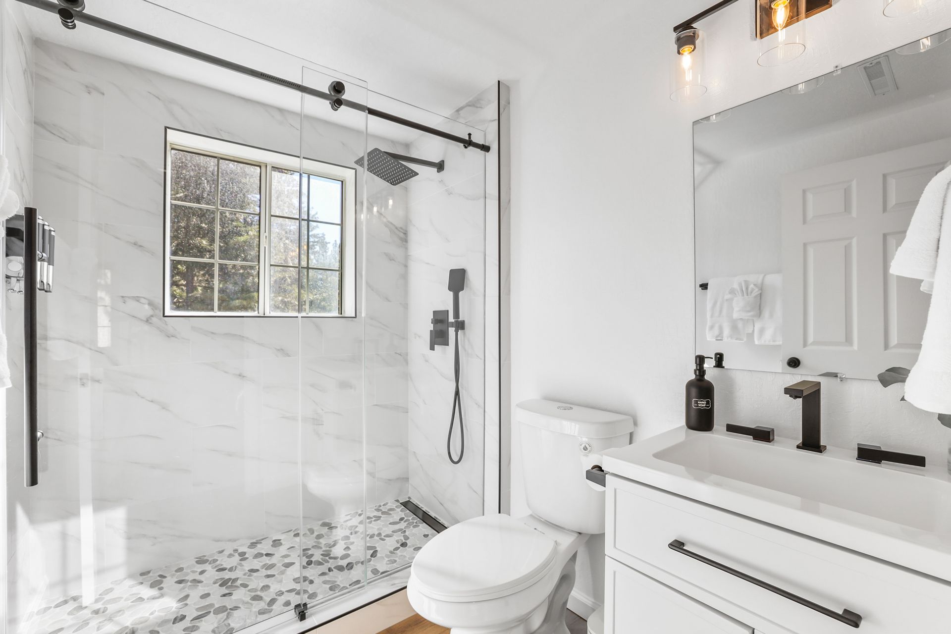 Bright white bathroom with glass shower, window, toilet, and vanity sink.