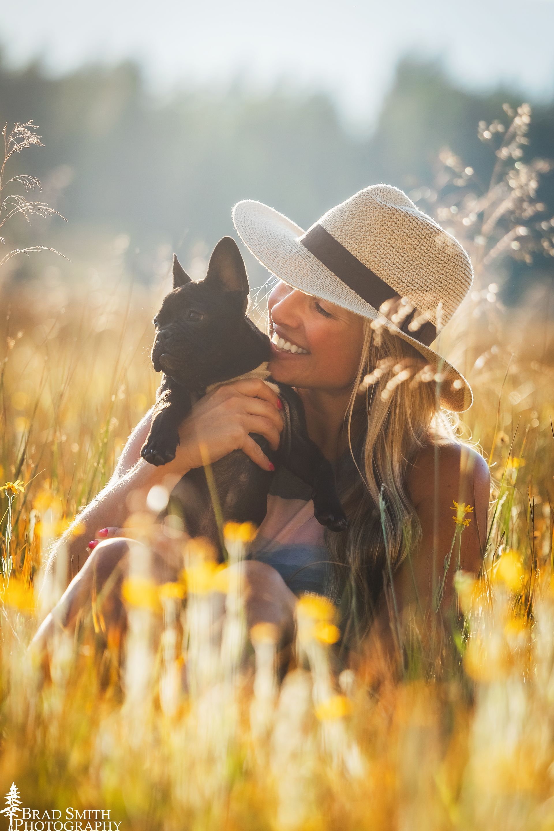 Person in a straw hat holding a black cat in a sunlit field of tall grass