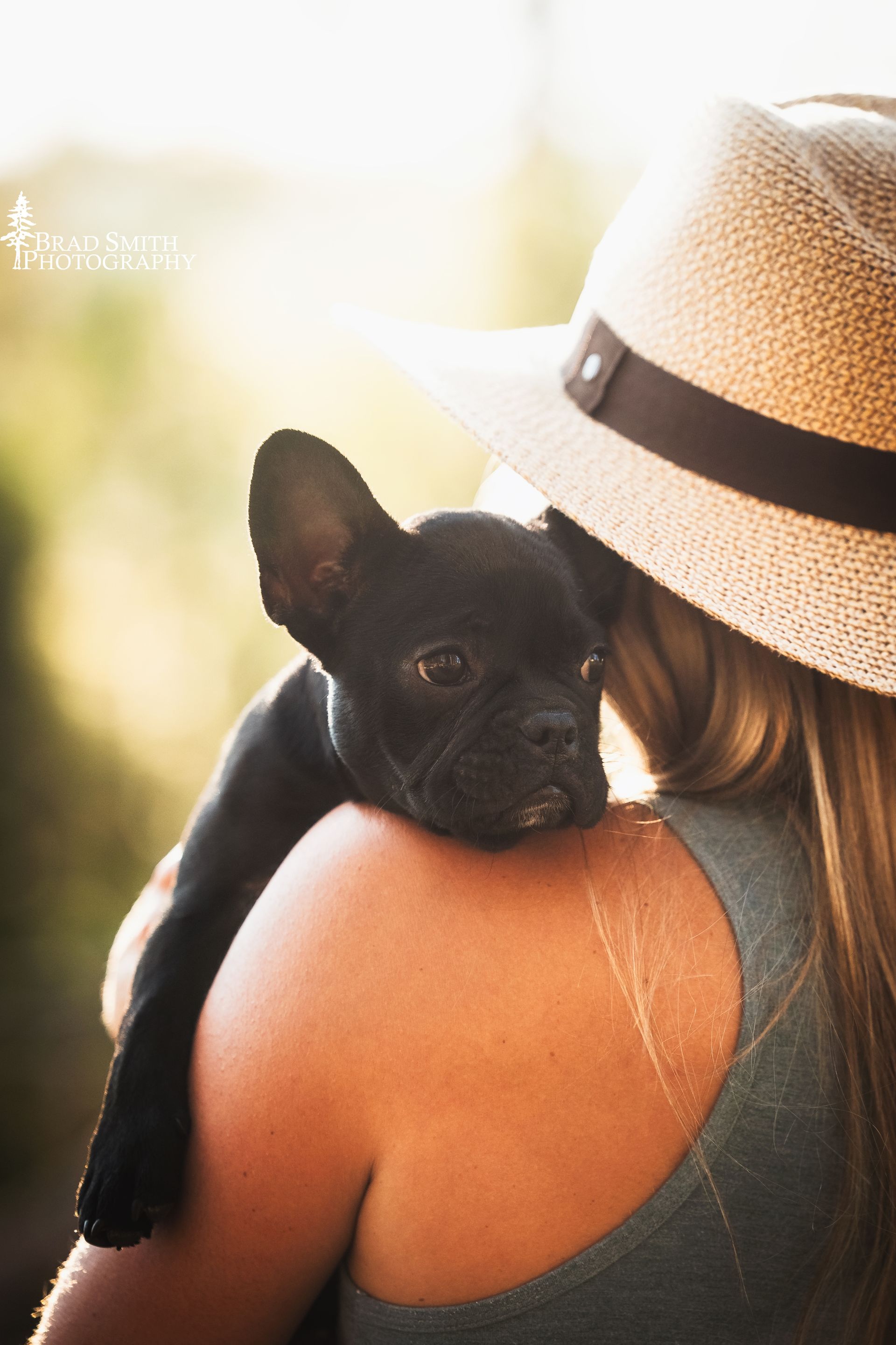 Black French bulldog resting on a woman's shoulder as she wears a straw hat outdoors