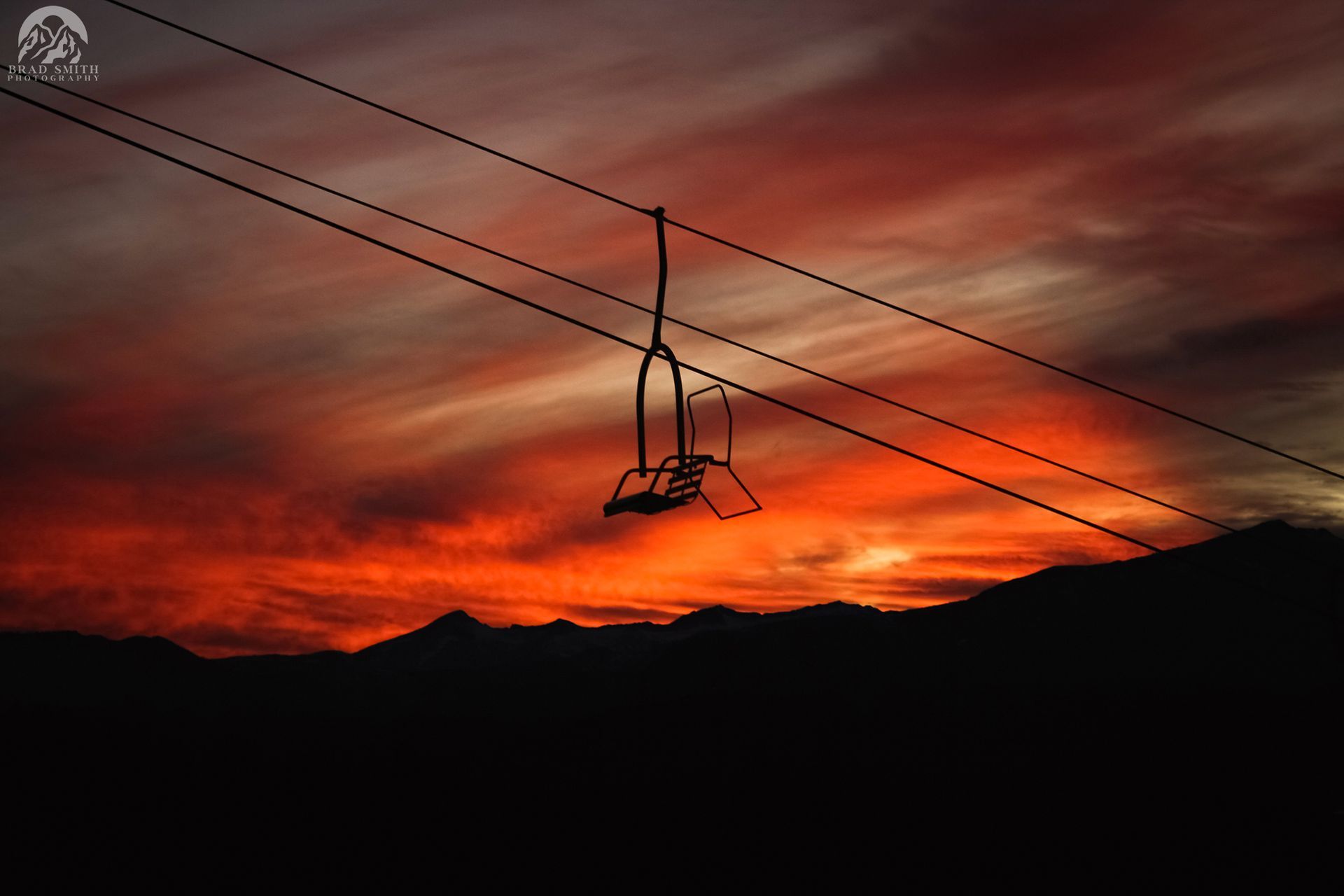 Silhouette of ski lift cables at sunset over dark mountains and a fiery orange sky
