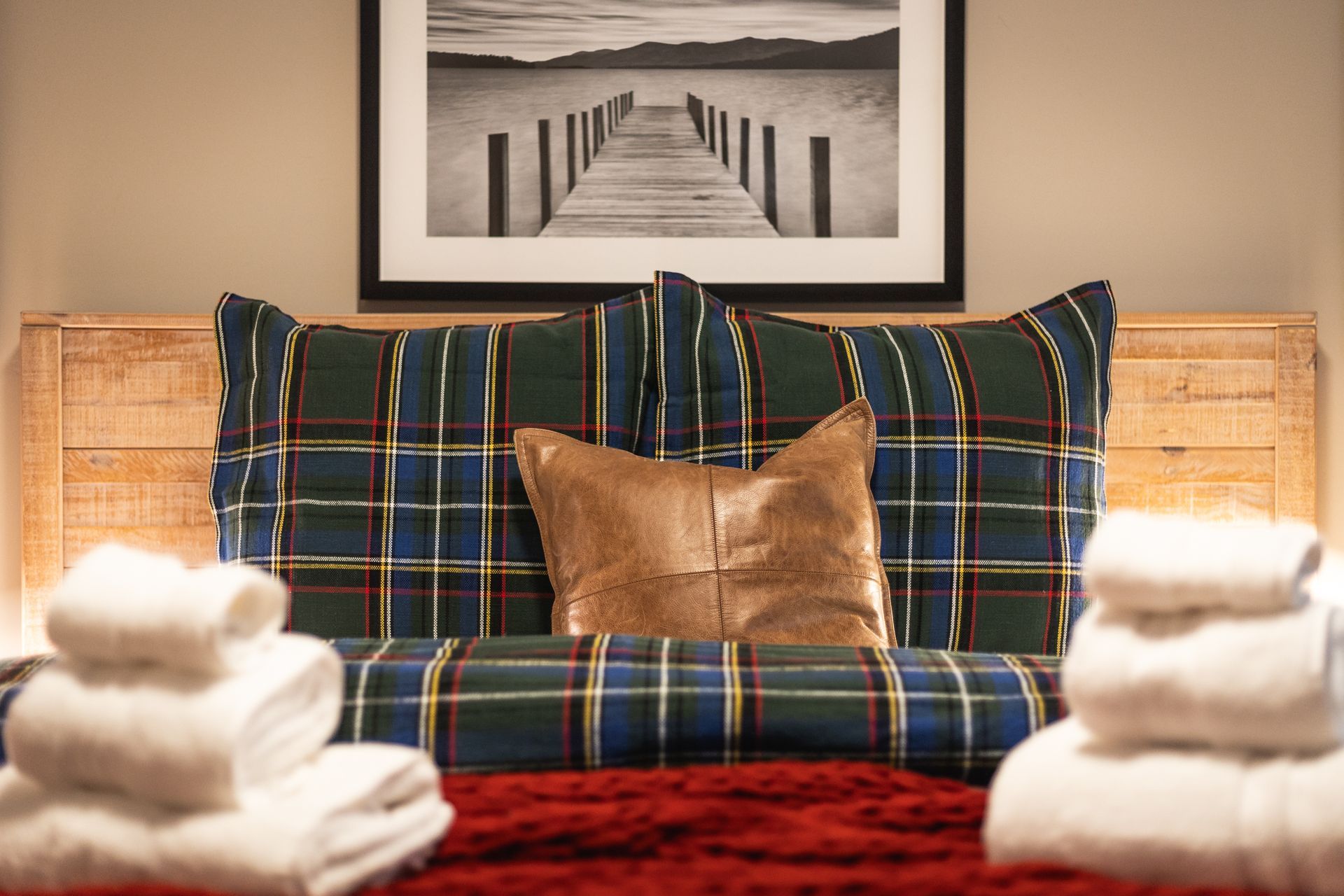 Plaid pillows on a woven bench with folded white towels, red throw, and a black-and-white pier print above