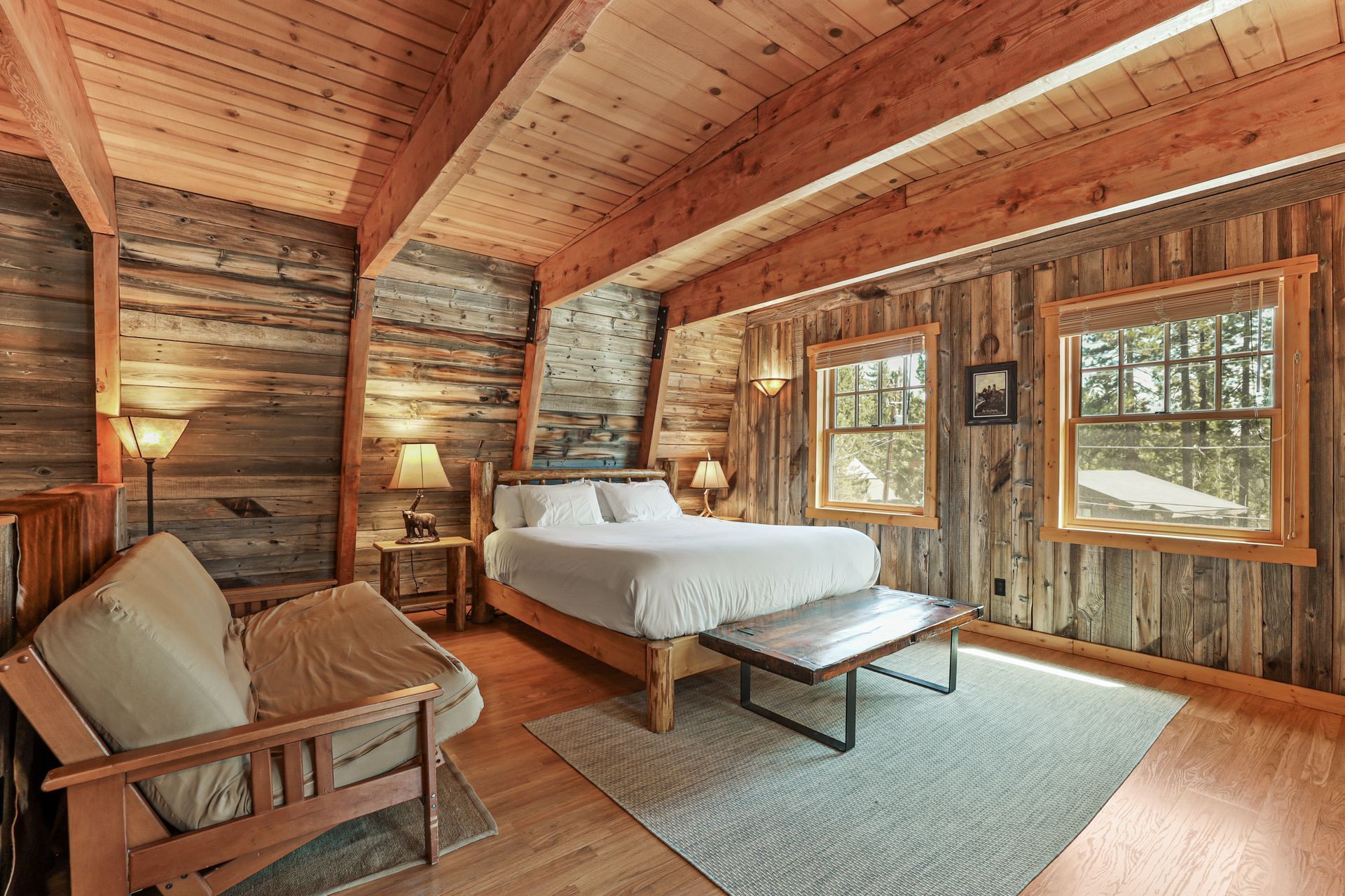 Cozy rustic cabin bedroom with wood-paneled walls, a white bed, sofa, and large windows