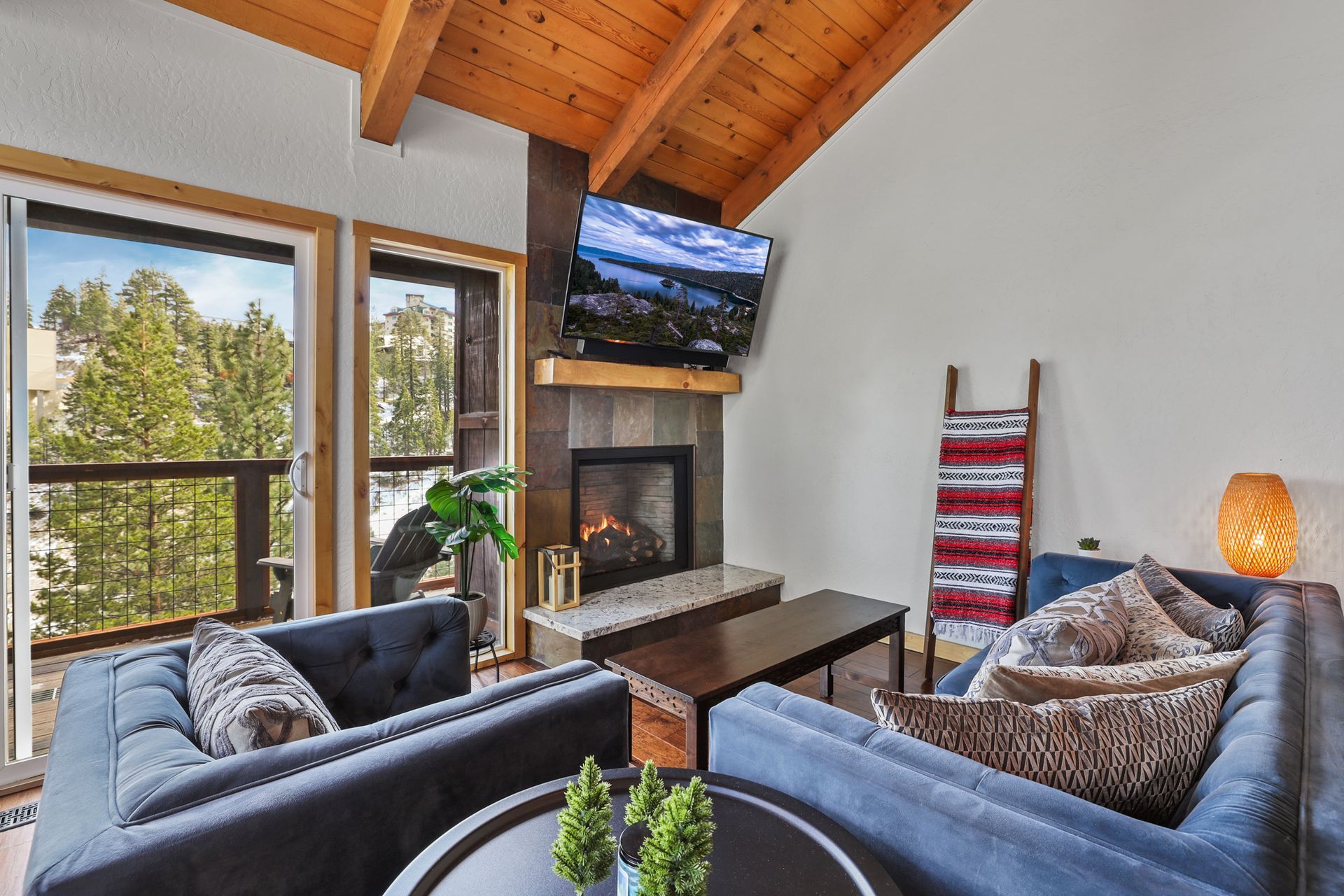 Cozy living room with fireplace, wall-mounted TV, blue chairs, and large windows overlooking trees