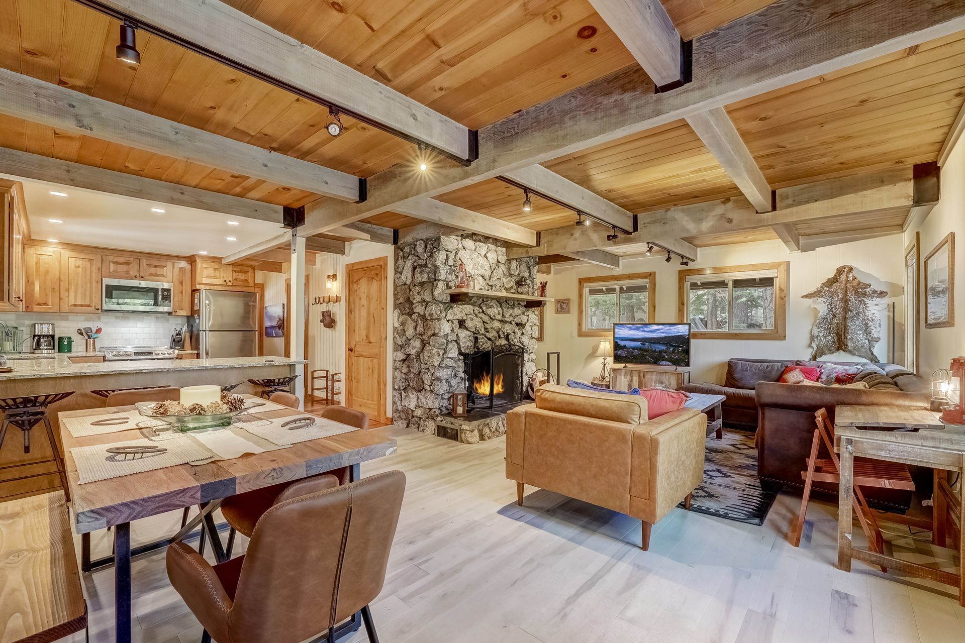 Cozy rustic living room with stone fireplace, wood beam ceiling, and open kitchen area