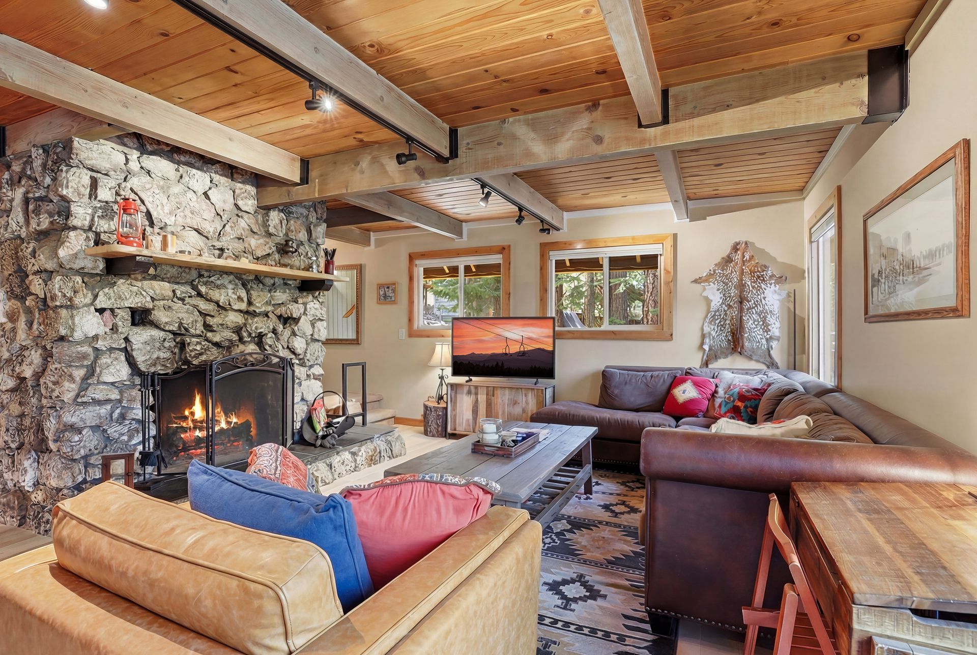 Cozy living room with stone fireplace, wood-beamed ceiling, and tan sectional sofa with colorful pillows