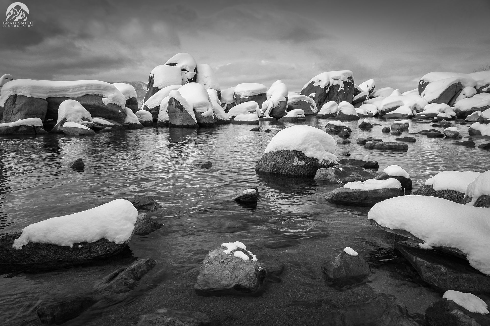 Snow-covered rocks in a shallow stream under a cloudy sky, in black and white.