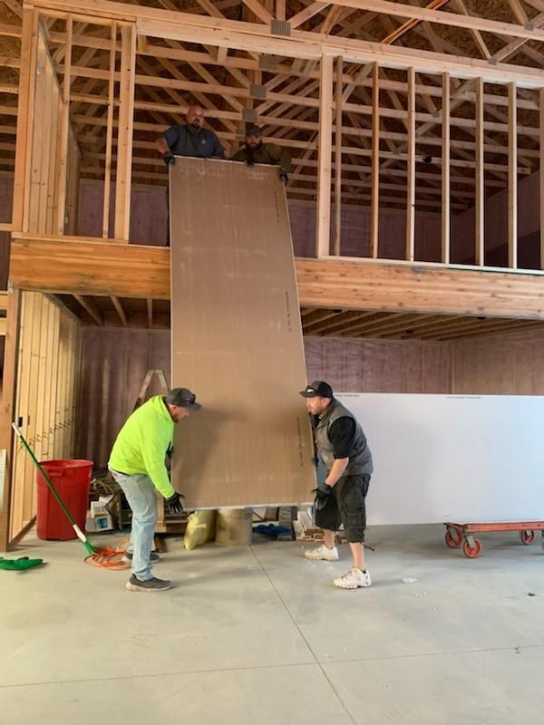 Two workers lift drywall, another on the loft. Inside a building under construction; concrete floor, wood framing.