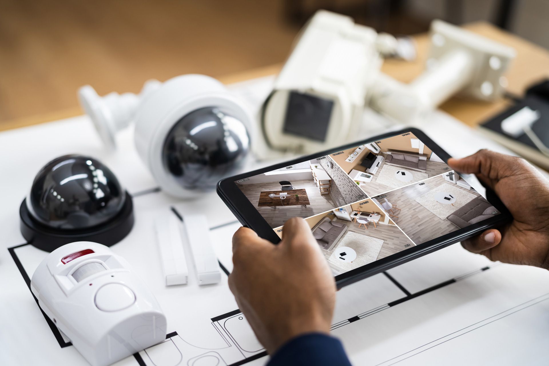 A person holds a tablet showing different security feeds above a table with various monitored alarm systems