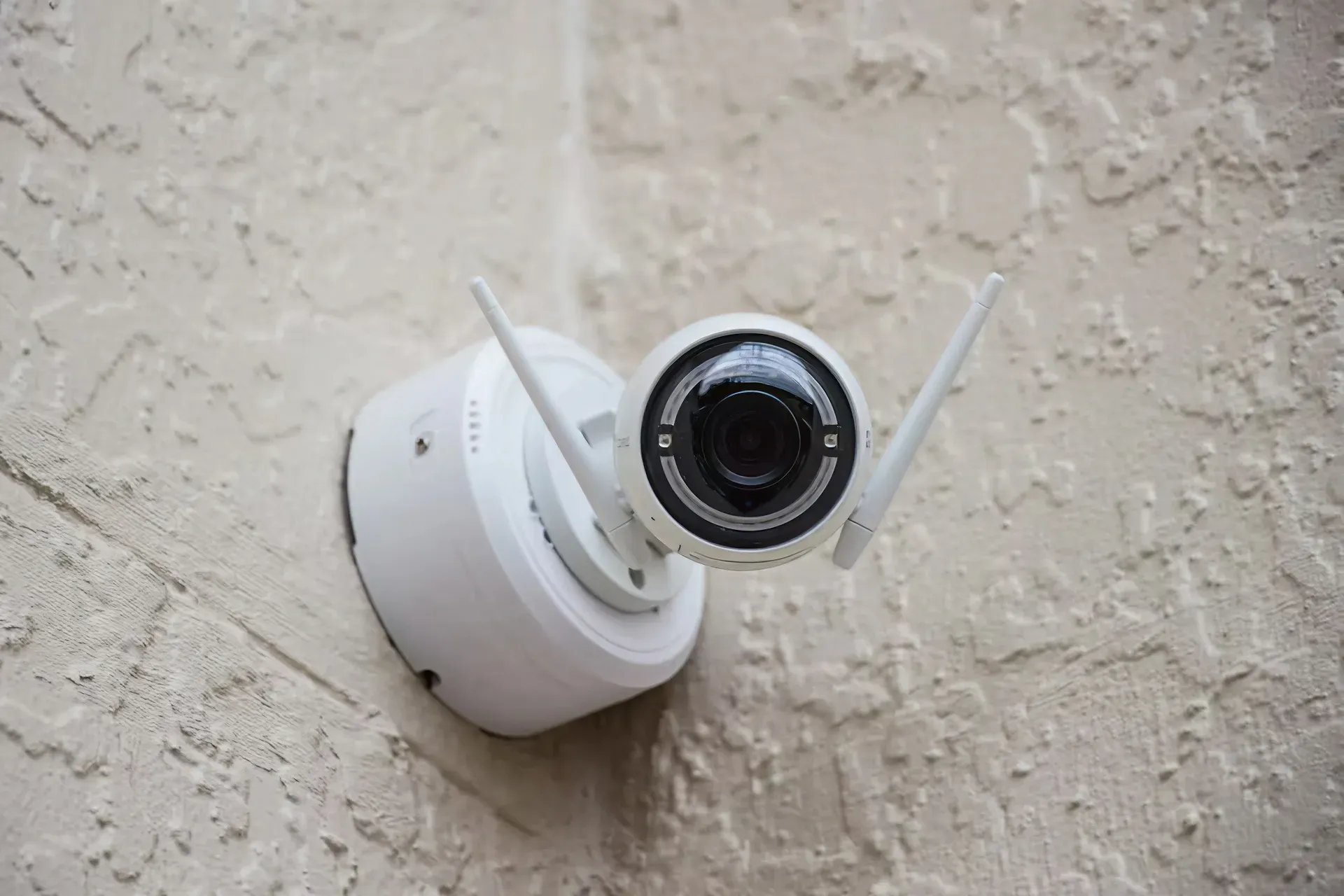 A white security camera is hoisted on a stucco wall