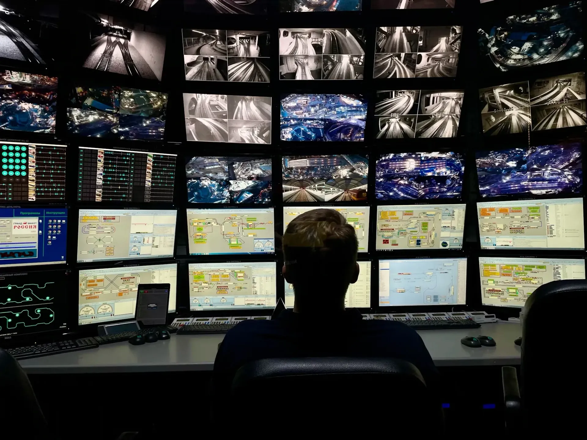 A man sits at a desk in front of multiple screens, watching security camera footage. 